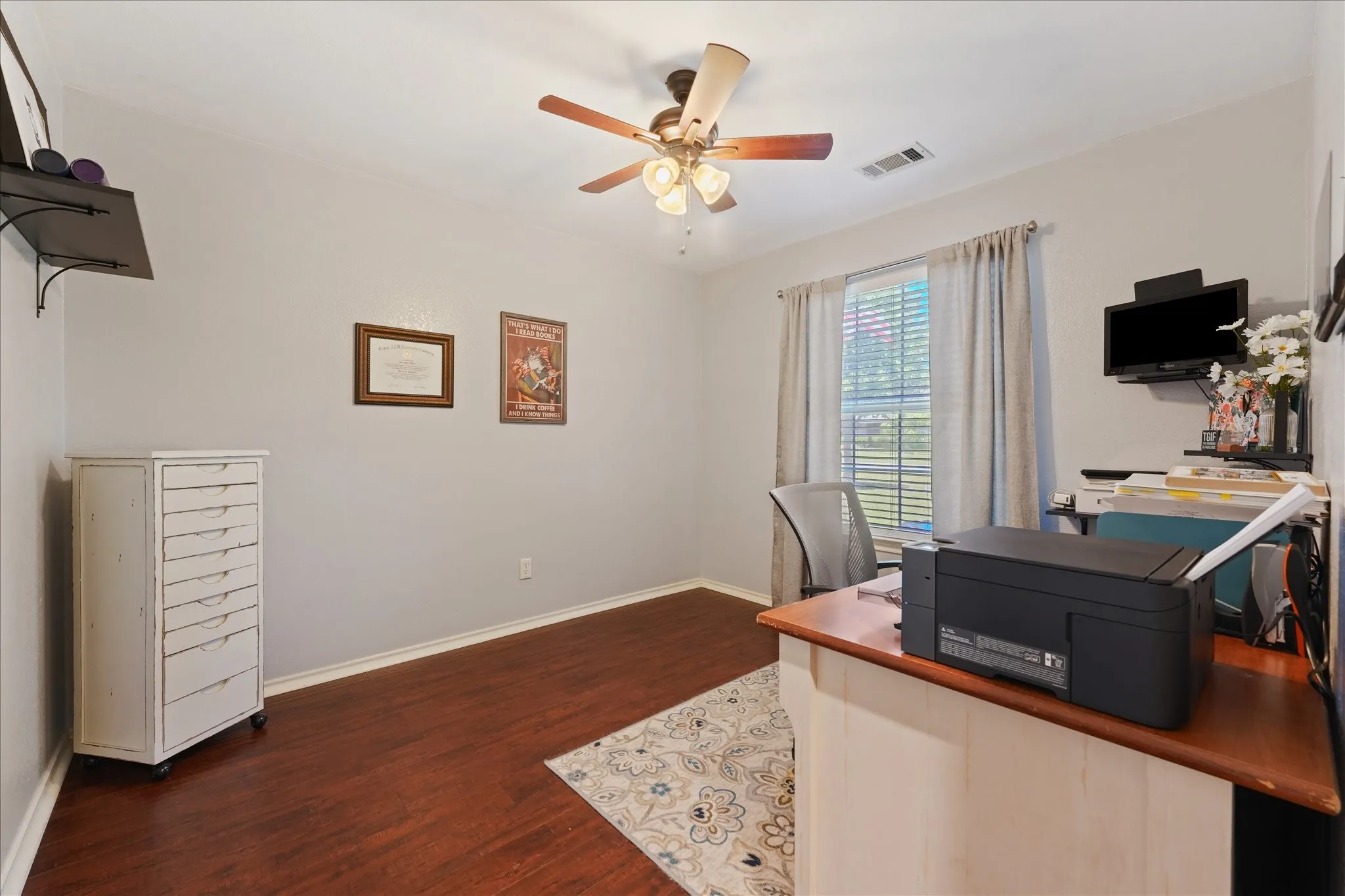 Bedroom being used as an office space featuring wood-finish flooring, a ceiling fan with integrated lighting, and a window with blinds and drapes