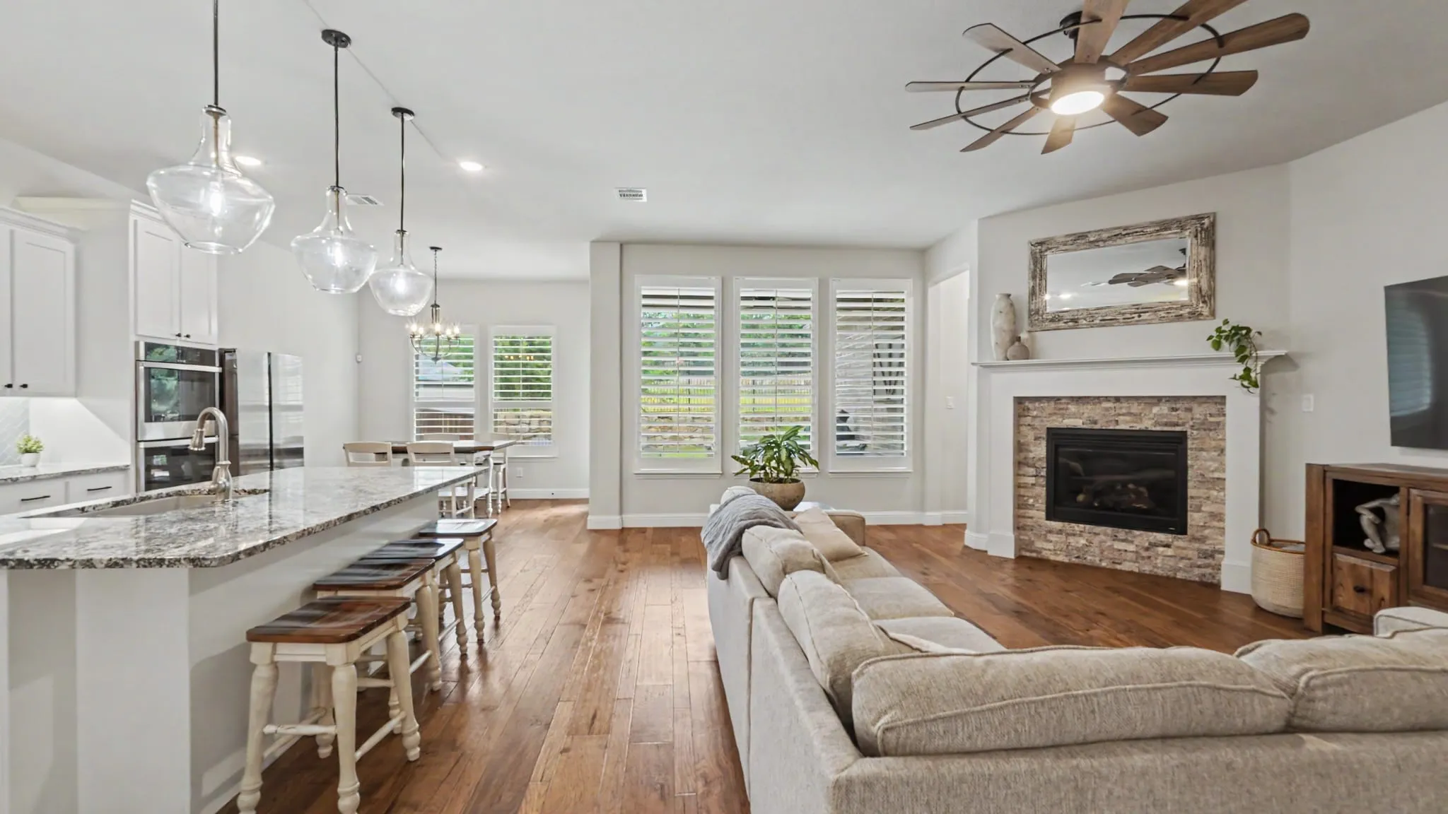 Open-concept living space featuring hardwood floors, a stone-surround fireplace with a white mantel, and a kitchen island with pendant lighting. Windows overlook backyard from the dining area, living, and kitchen.