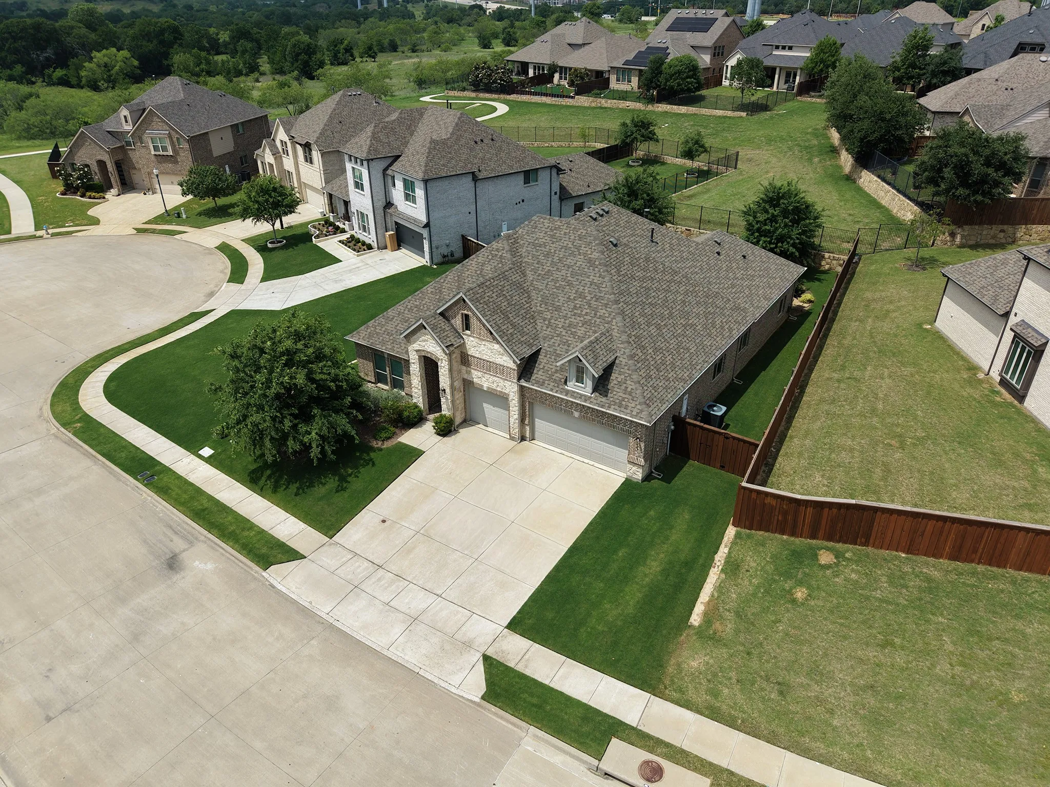 Aerial view of single-story residence featuring a three-car garage, stone and brick facade, gabled roofline, and established front landscaping and naturally green yard (not edited).