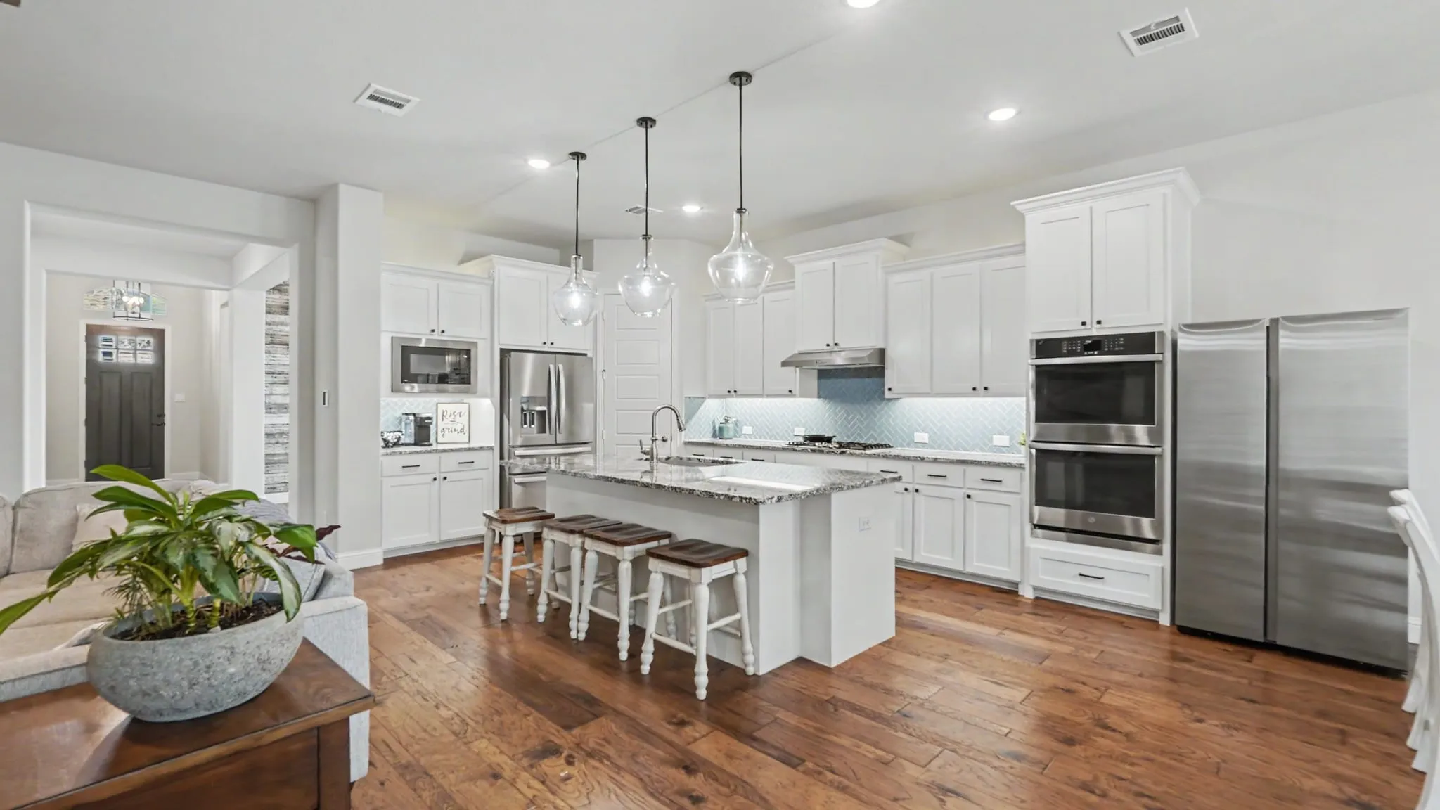 Open-concept kitchen featuring hardwood floors, white cabinetry, a central island with bar seating, pendant lighting, and stainless steel appliances including double ovens and a two side-by-side refrigerators.