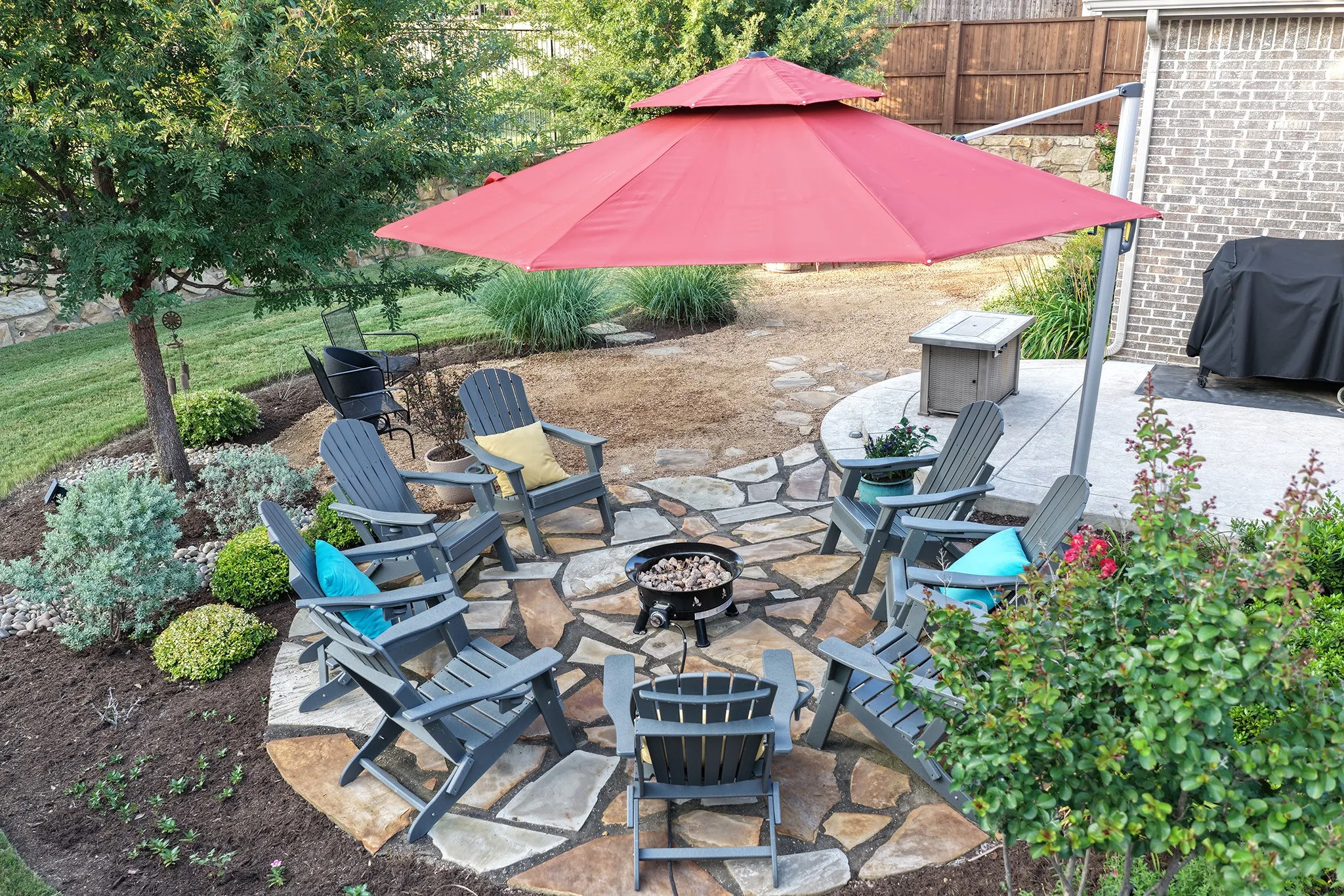 Outdoor flagstone patio featuring a central fire pit area, surrounded by lush landscaping and mature trees.