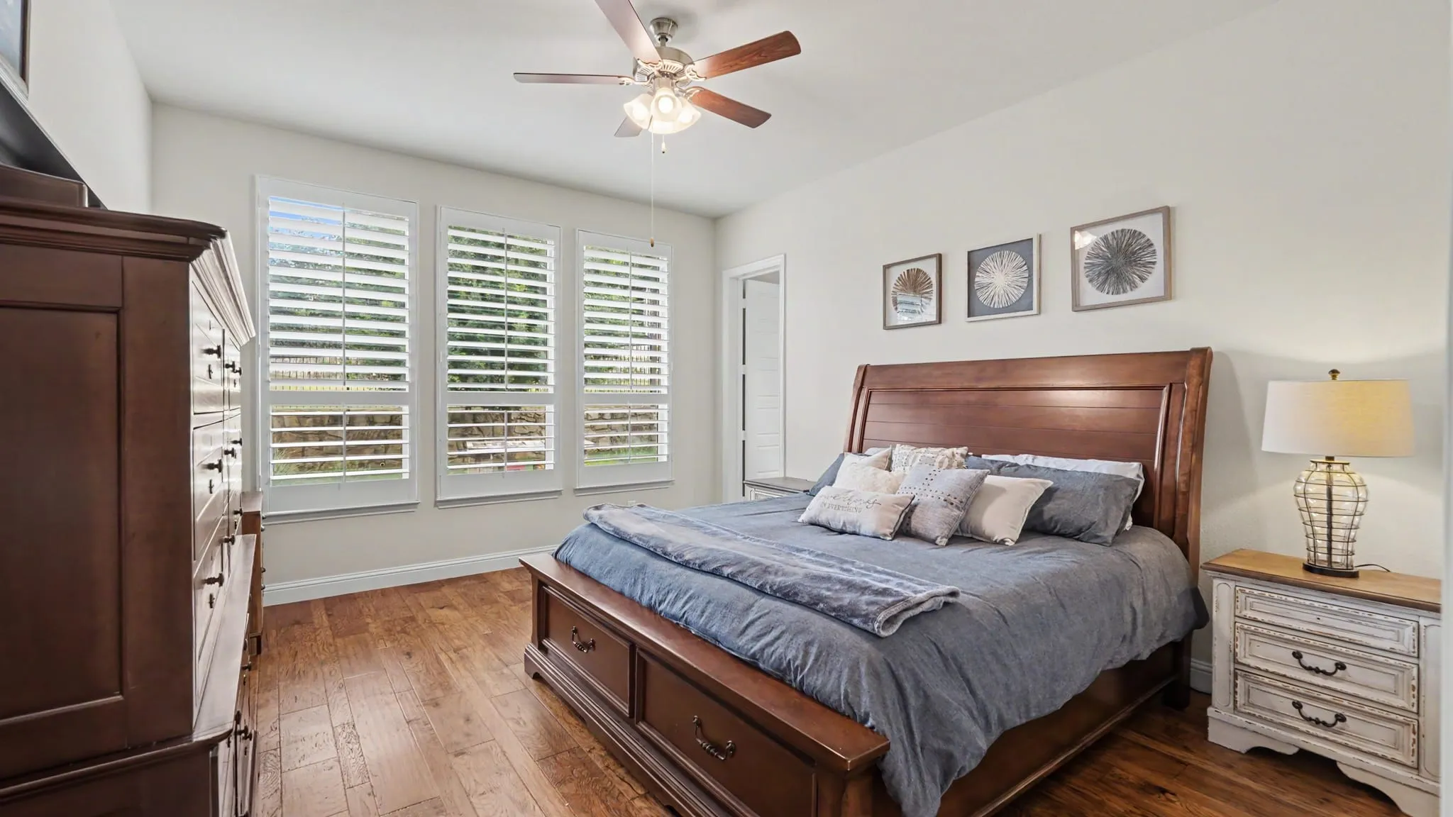 Bedroom featuring hardwood floors, three windows with white plantation shutters, and a ceiling fan with integrated lighting.