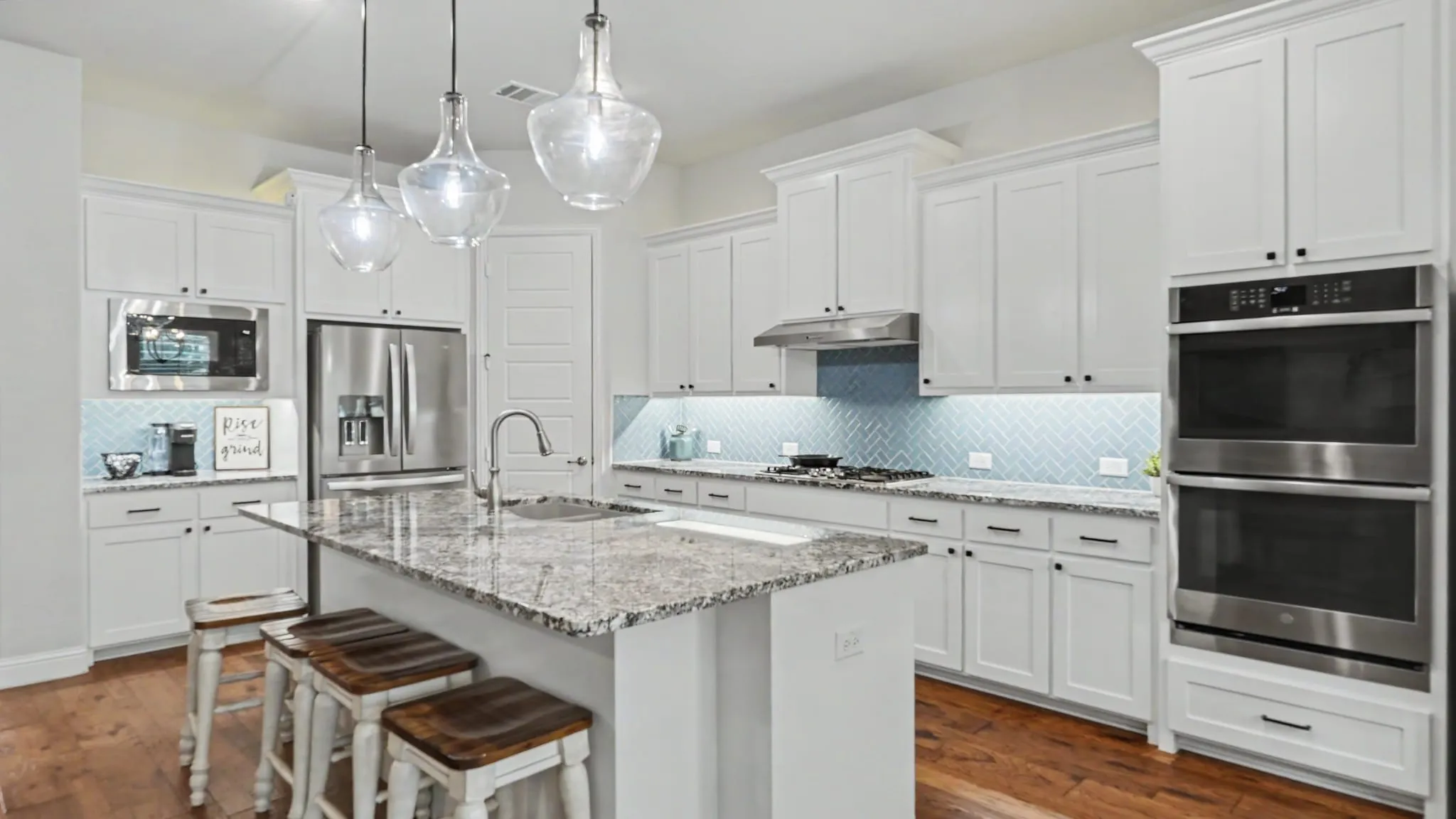 Spacious kitchen featuring an expansive central island with a granite countertop and undermount sink, white shaker-style cabinetry, stainless steel appliances including double wall ovens, a herringbone tile light blue backsplash, and hardwood floors.