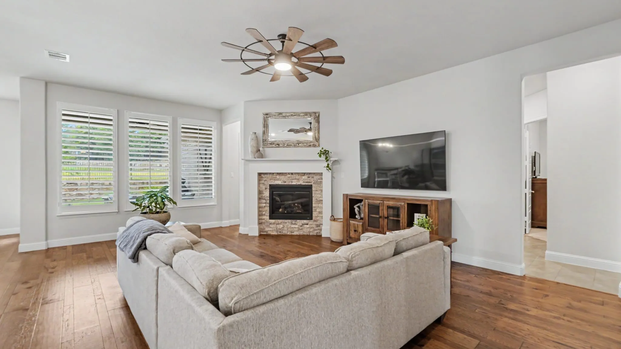 Living area featuring hardwood floors, a corner fireplace with a stone surround and white mantel, and a ceiling fan with integrated lighting.  Plantation shutter windows overlook the backyard.