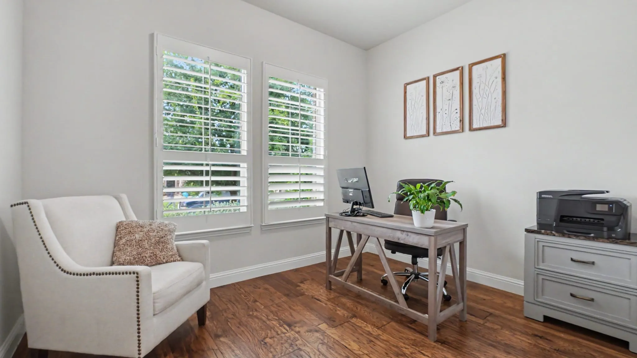 Front bedroom/office  featuring hardwood floors, two windows with white plantation shutters that overlook the front yard shaded by a tree, and light-toned wall paint.