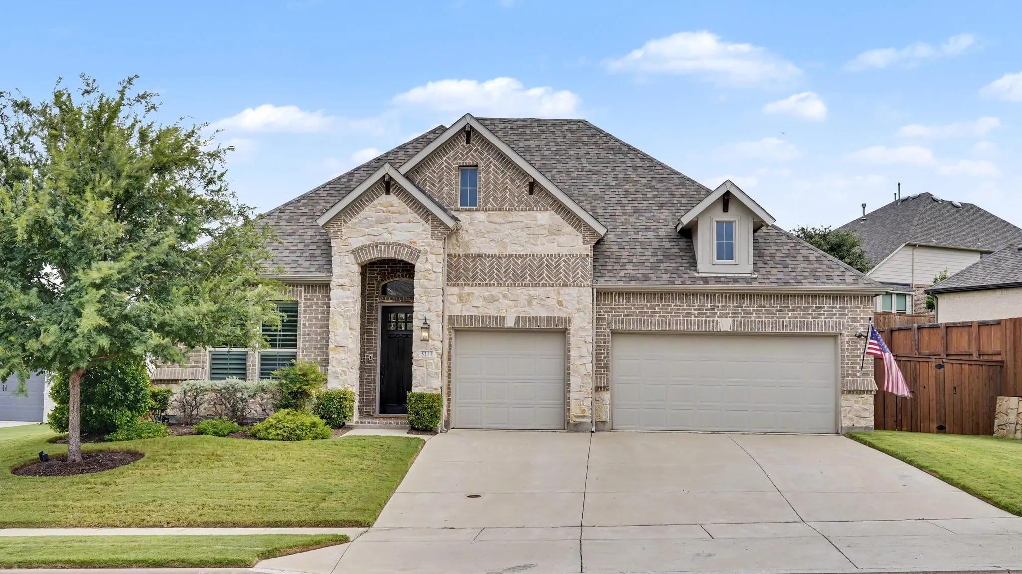 Front of home showcasing three-car garage and beautiful tree, with pristine landscaping.