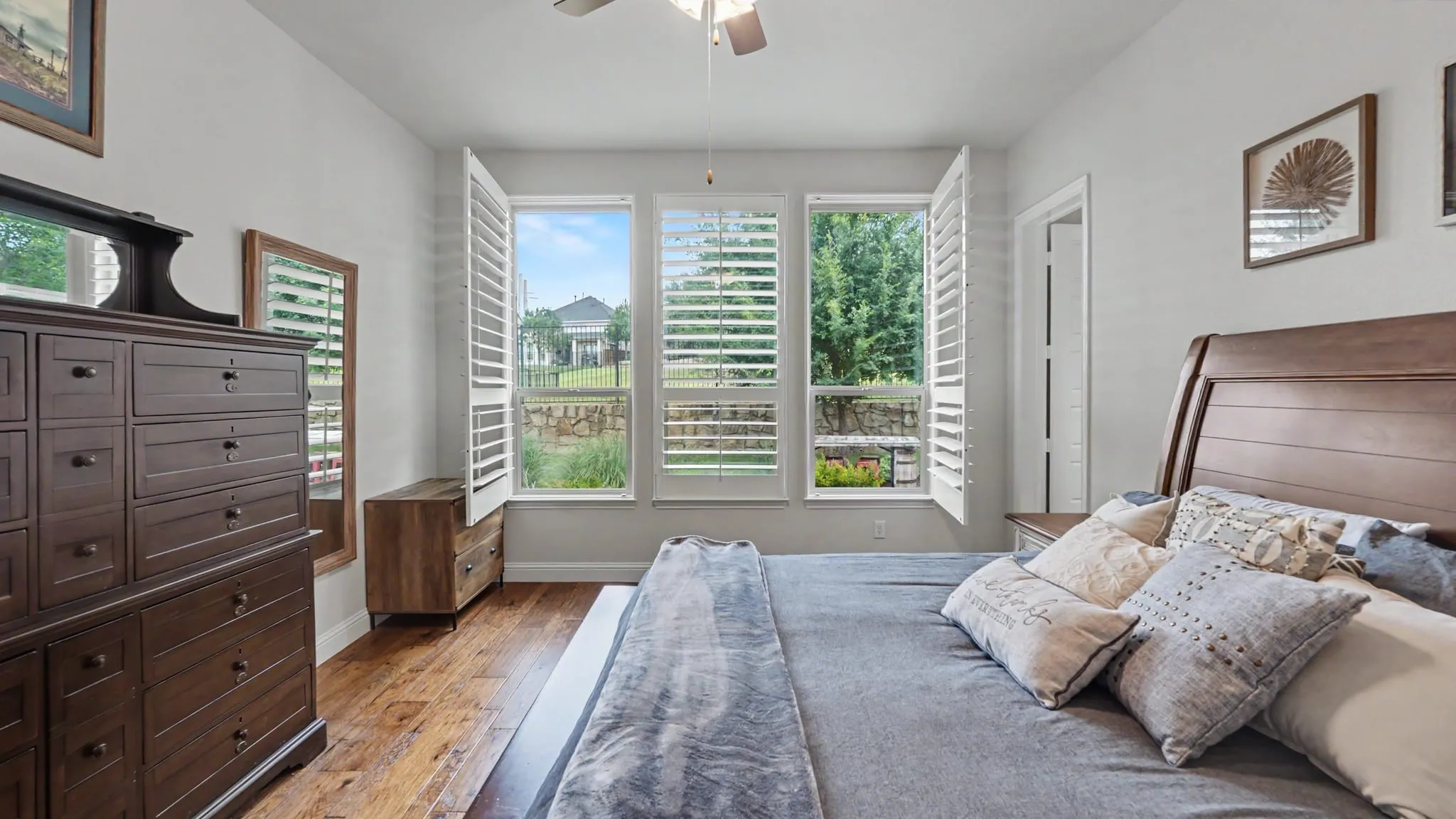 Bedroom featuring hardwood floors, three windows with white plantation shutters, and a ceiling fan with integrated lighting.