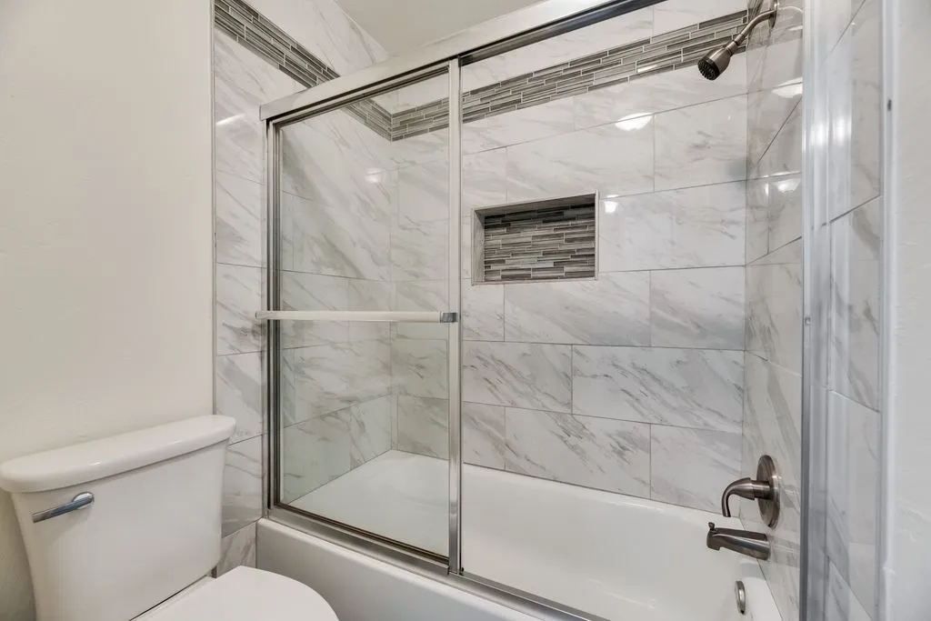 Bathtub and shower combination featuring a glass sliding door enclosure, large format white tile surround with subtle veining, and a decorative mosaic tile accent band