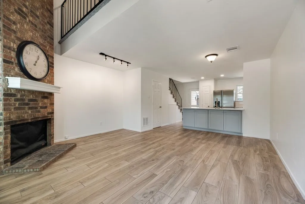 Spacious living area featuring a floor-to-ceiling brick fireplace with a white mantel, wood-finish flooring, and a modern kitchen island with light blue cabinetry and a light-toned countertop
