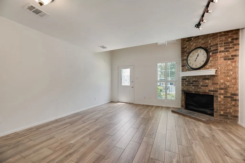 Spacious room featuring wood-finish flooring, a brick-faced fireplace with a white mantel, and a white paneled door