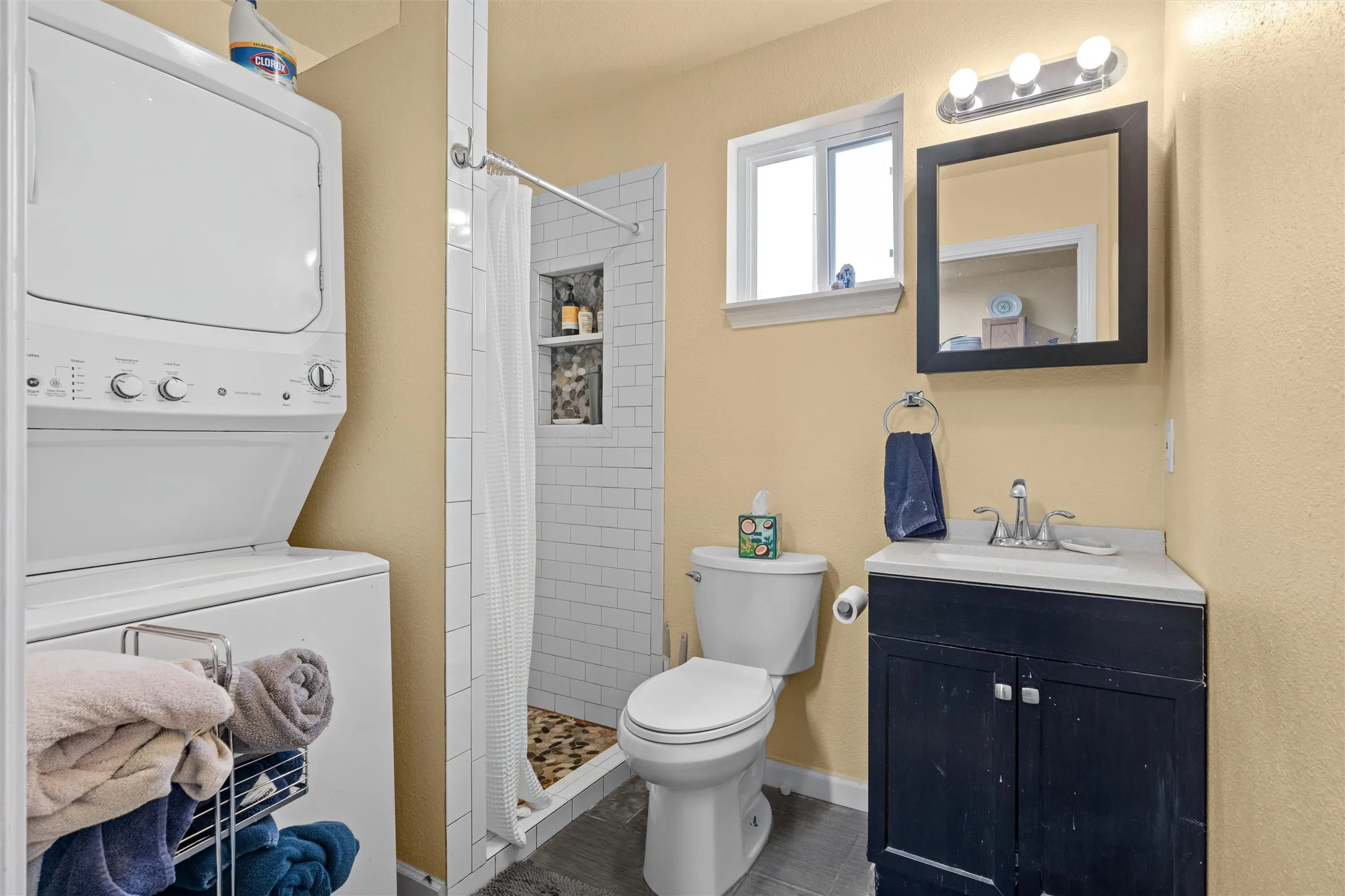 Bathroom featuring a stacked laundry unit, a walk-in shower with tiled walls and a stone pebble floor, a single window, a dark-toned vanity with a white countertop, and a wall-mounted mirror with overhead lighting