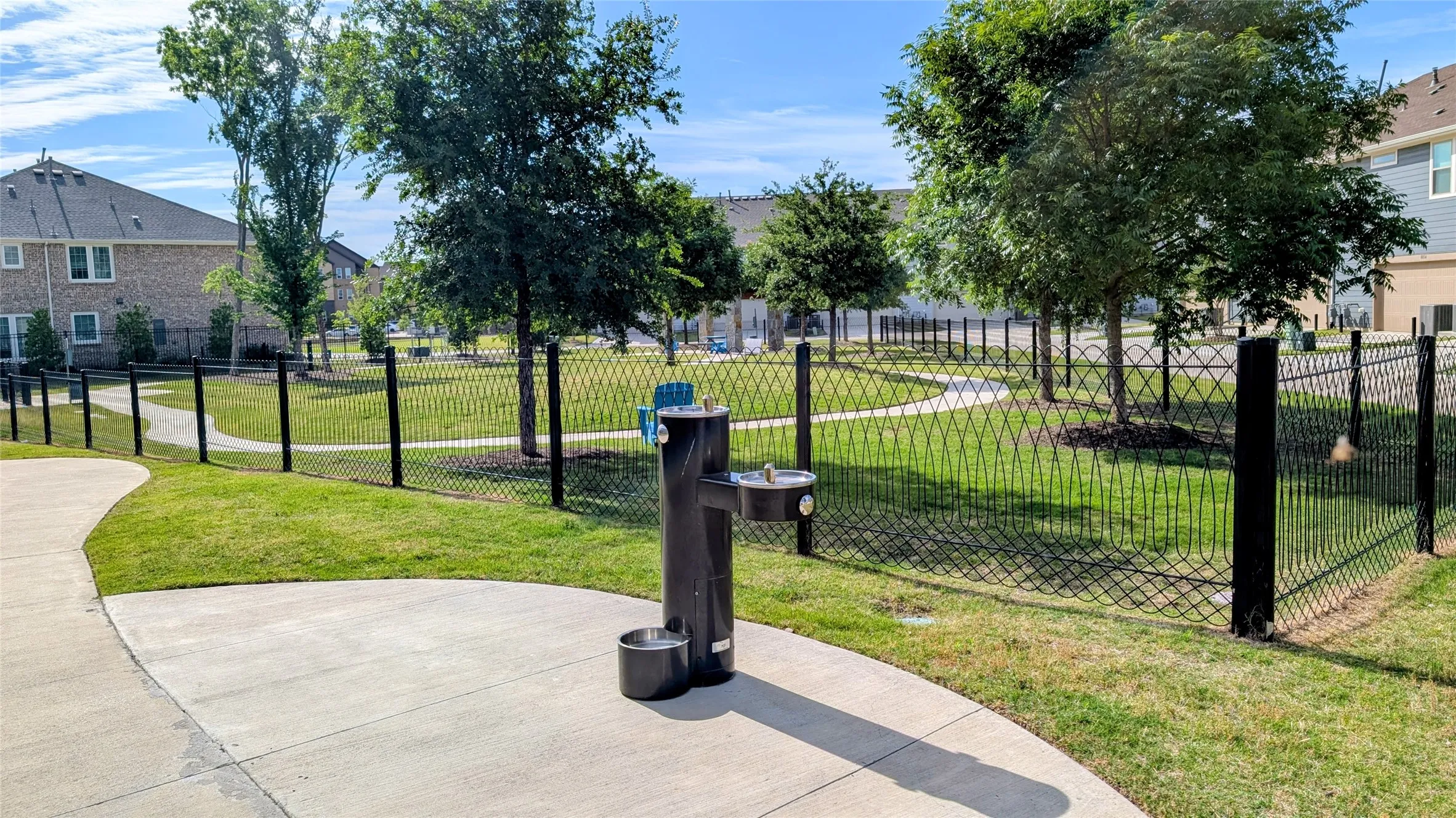 Community green space featuring a paved walkway, mature trees, and a black wrought iron fence