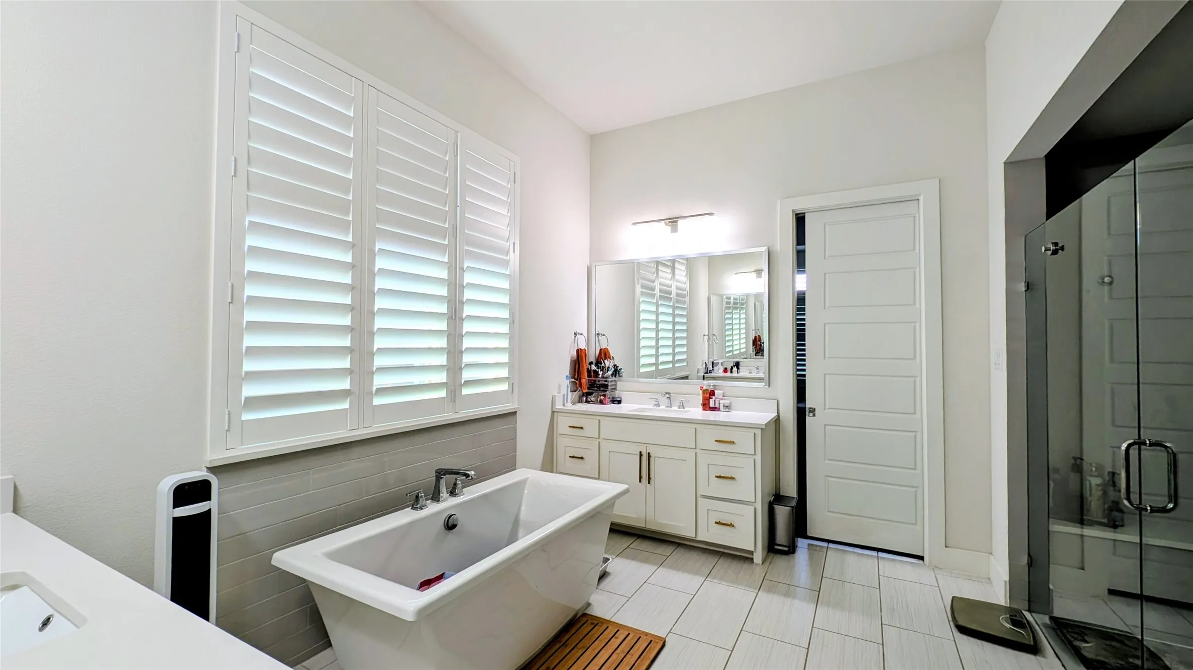 Freestanding soaking tub with a tiled surround, plantation shutters, dual vanity with undermount sinks, and a frameless glass shower enclosure