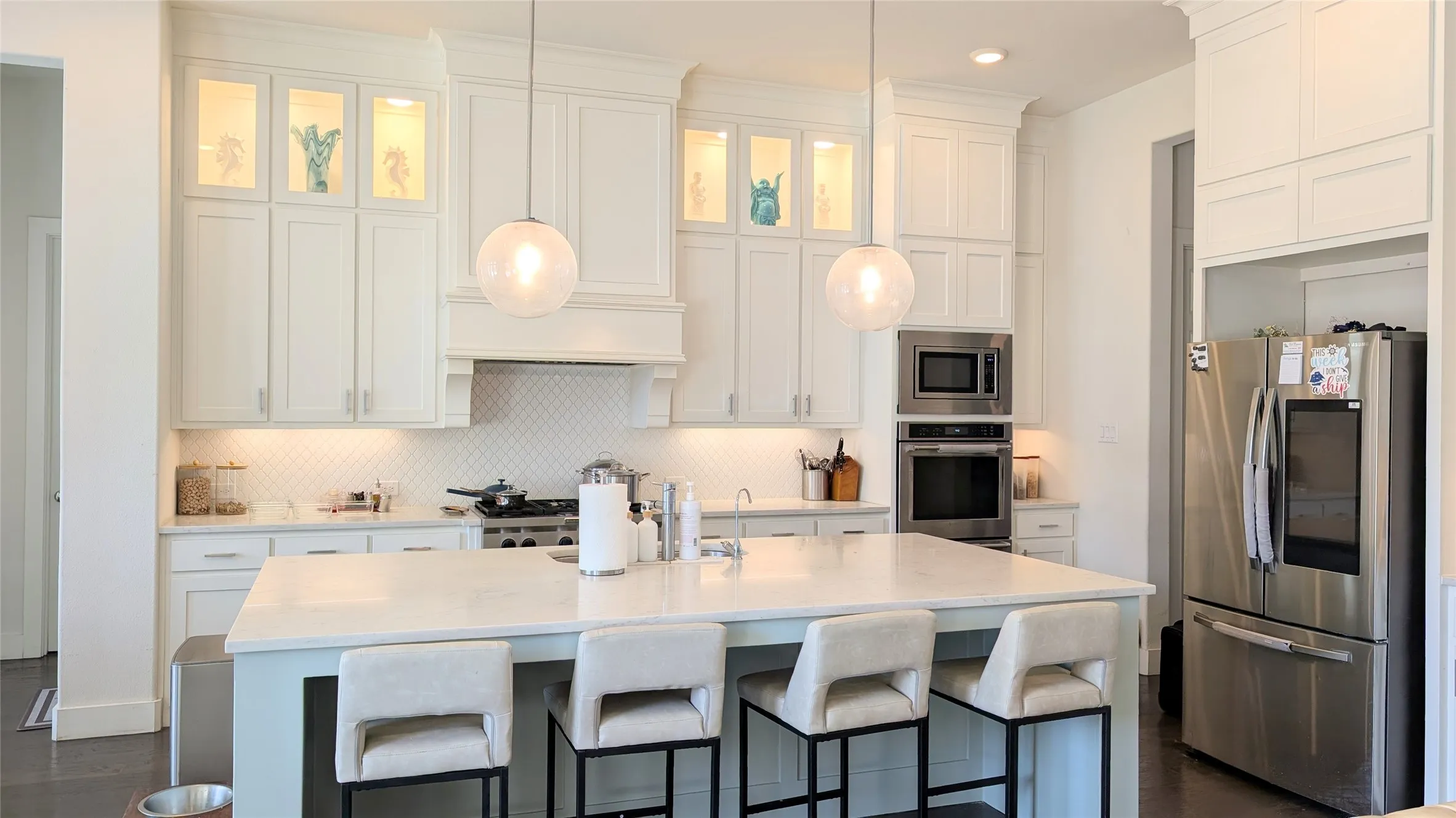 Gourmet kitchen featuring a large central island with seating, stainless steel appliances, white shaker cabinetry with crown molding, glass-front upper cabinets, and a white tile backsplash