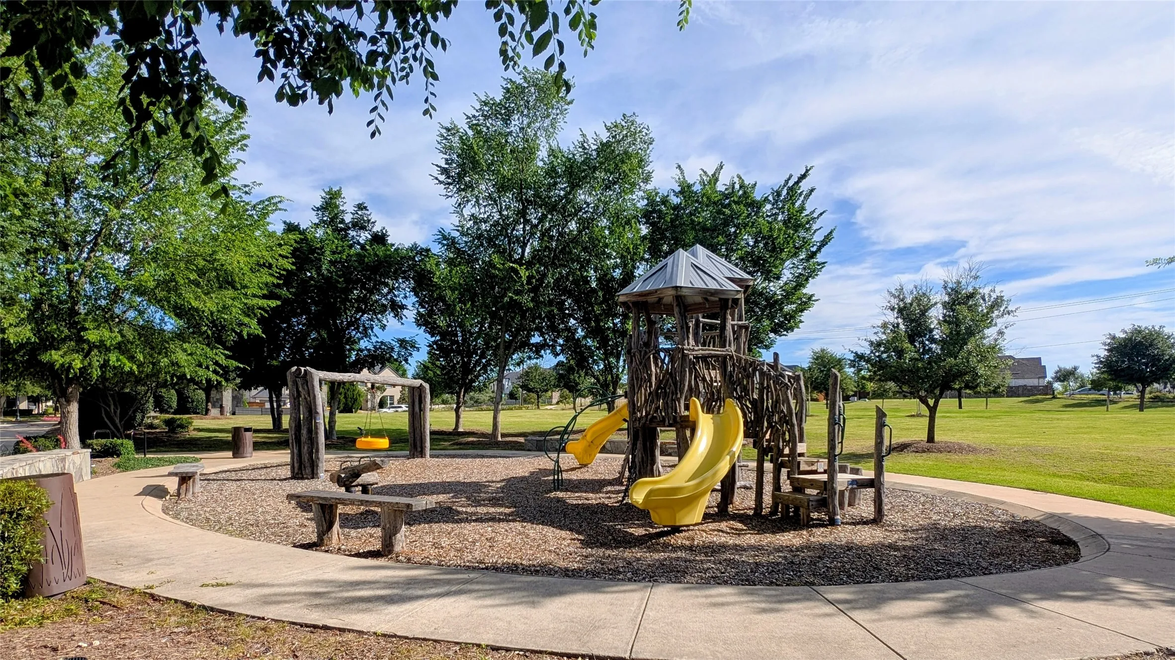 Community playground featuring a wood-look play structure with two yellow slides, a swing set, and integrated seating areas
