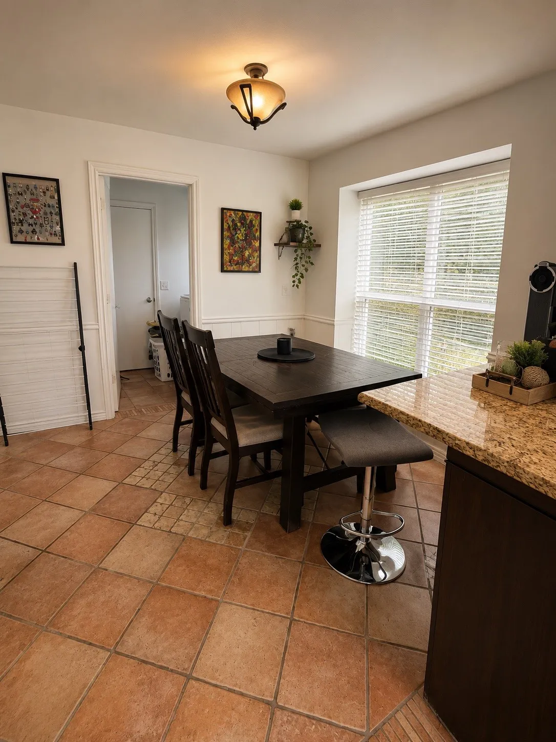 Dining area featuring terracotta-style floor tiling, a large window with blinds, and a built-in kitchen peninsula with a granite-style countertop