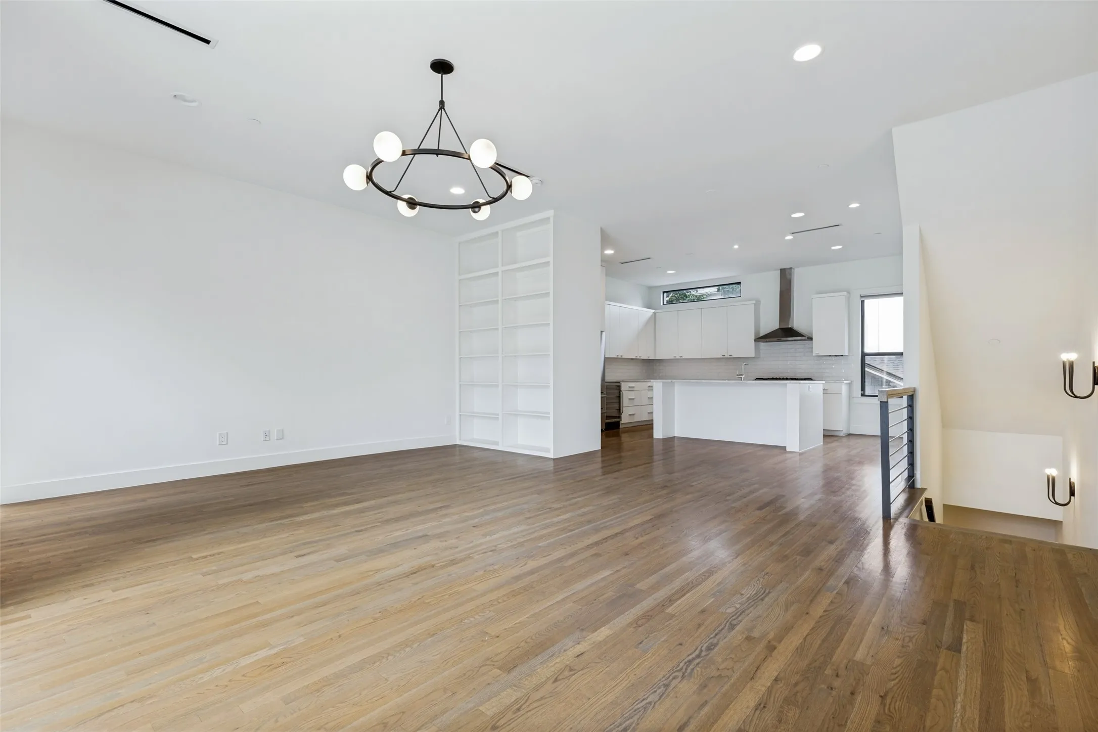 Open-concept living space featuring wood-finish flooring, a contemporary chandelier, built-in shelving, a kitchen with white cabinetry, and a staircase with a modern railing