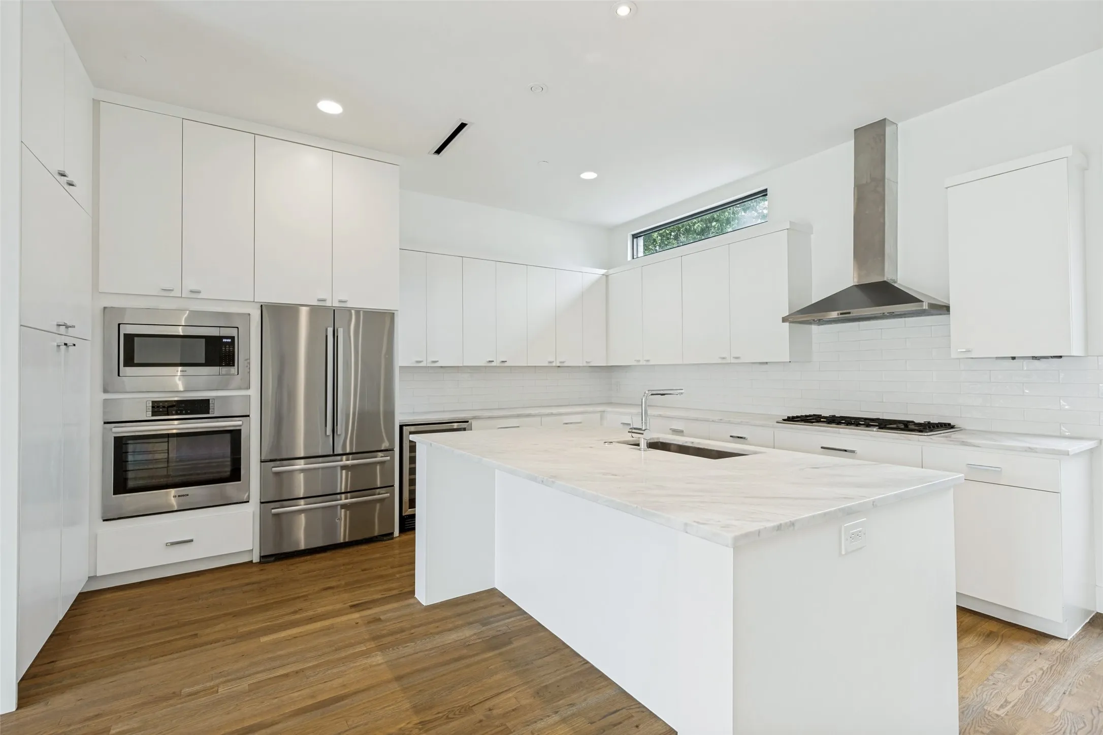 Contemporary kitchen featuring white cabinetry, a large center island with a light-toned countertop, stainless steel appliances, a range hood, and wood-finish flooring