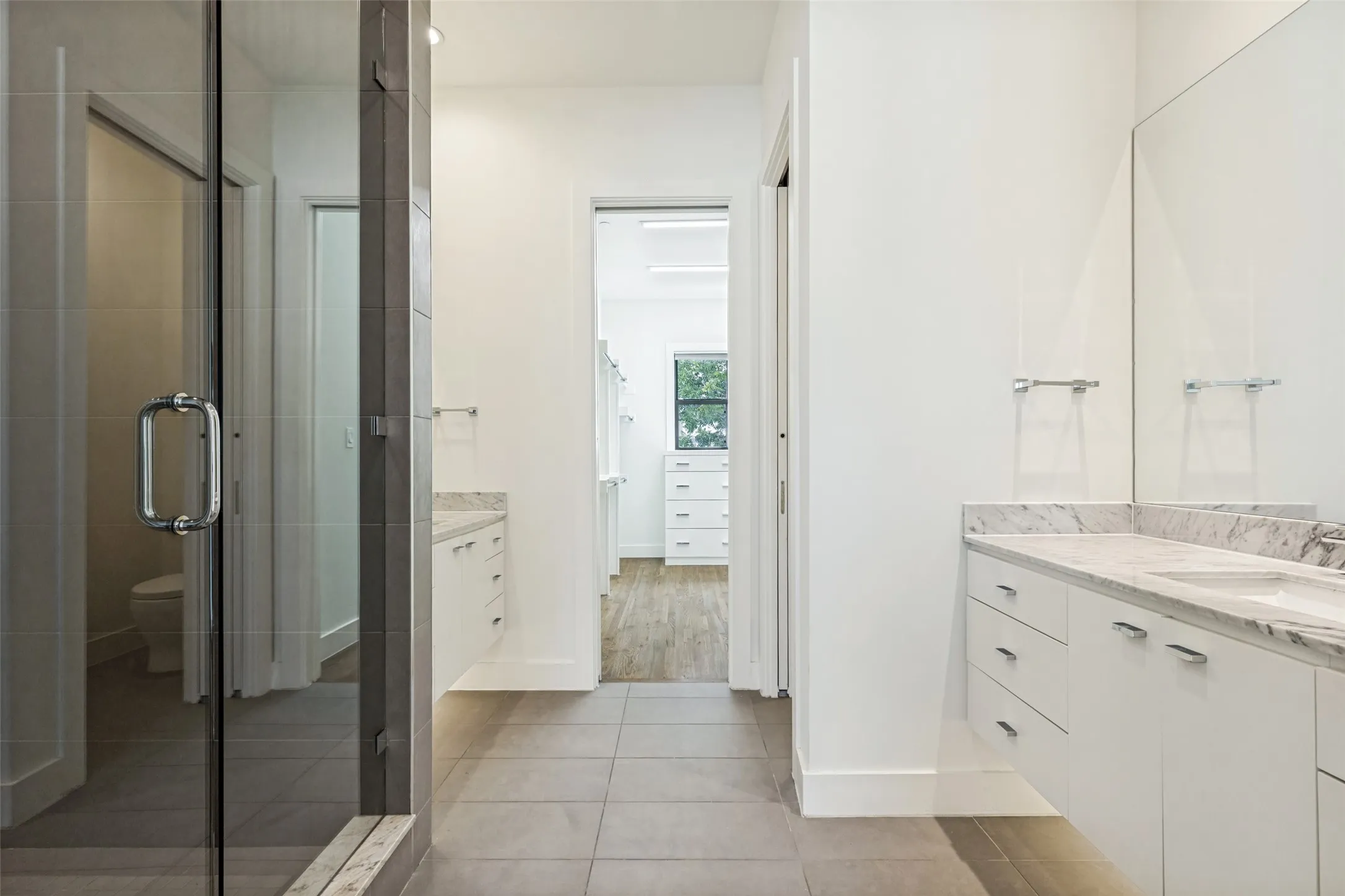 Modern bathroom featuring a frameless glass shower enclosure, dual white vanities with light-toned stone countertops, and large format tile flooring