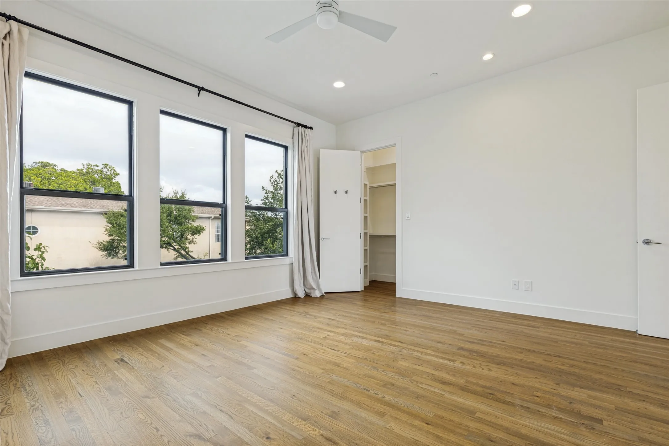 Spacious room featuring natural wood-finish flooring, a trio of large windows with black frames, recessed lighting, and a ceiling fan