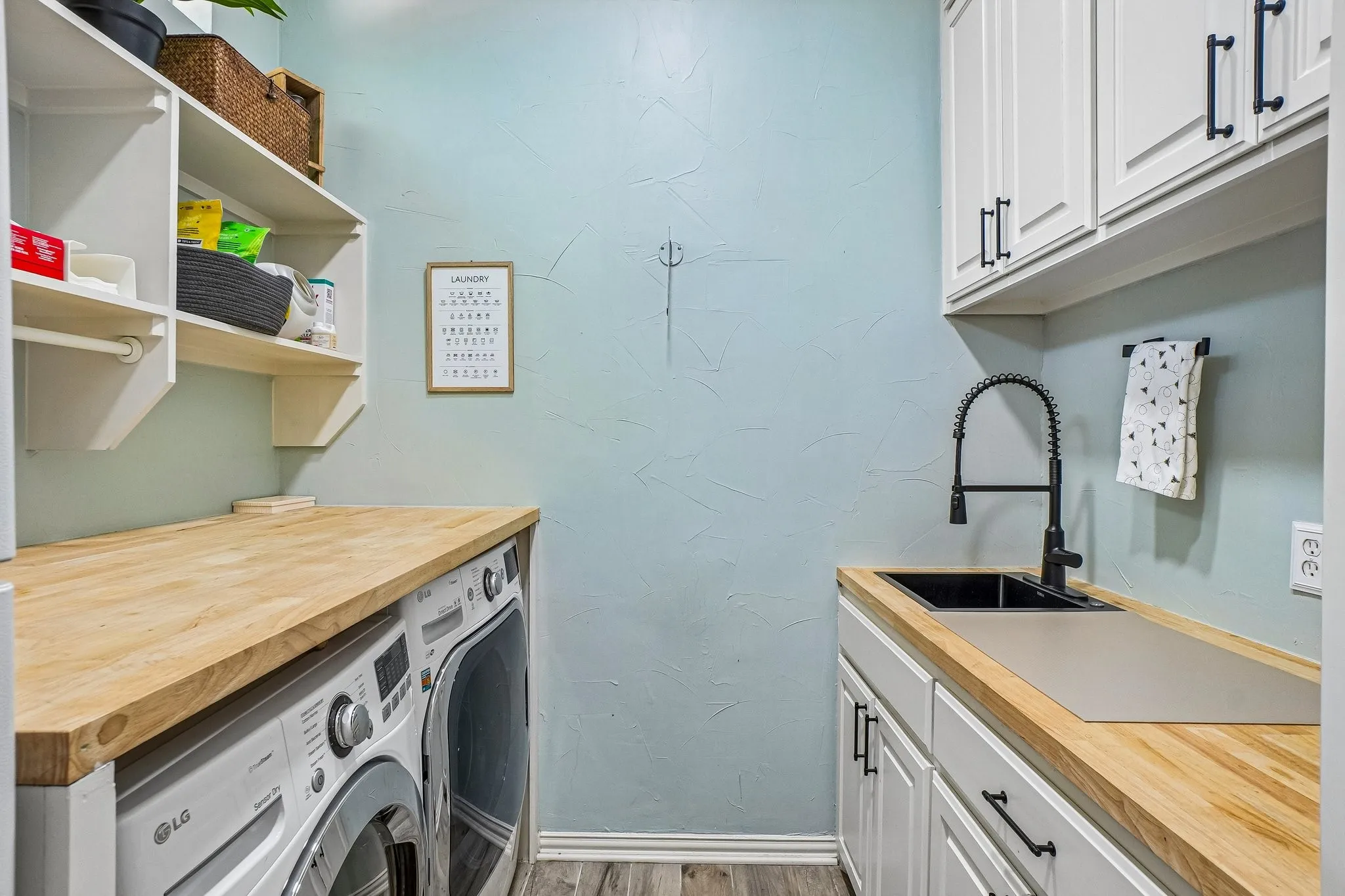 Dedicated laundry room featuring light wood-finish countertops, white shaker-style cabinetry with matte black hardware, and a black gooseneck faucet with an undermount sink
