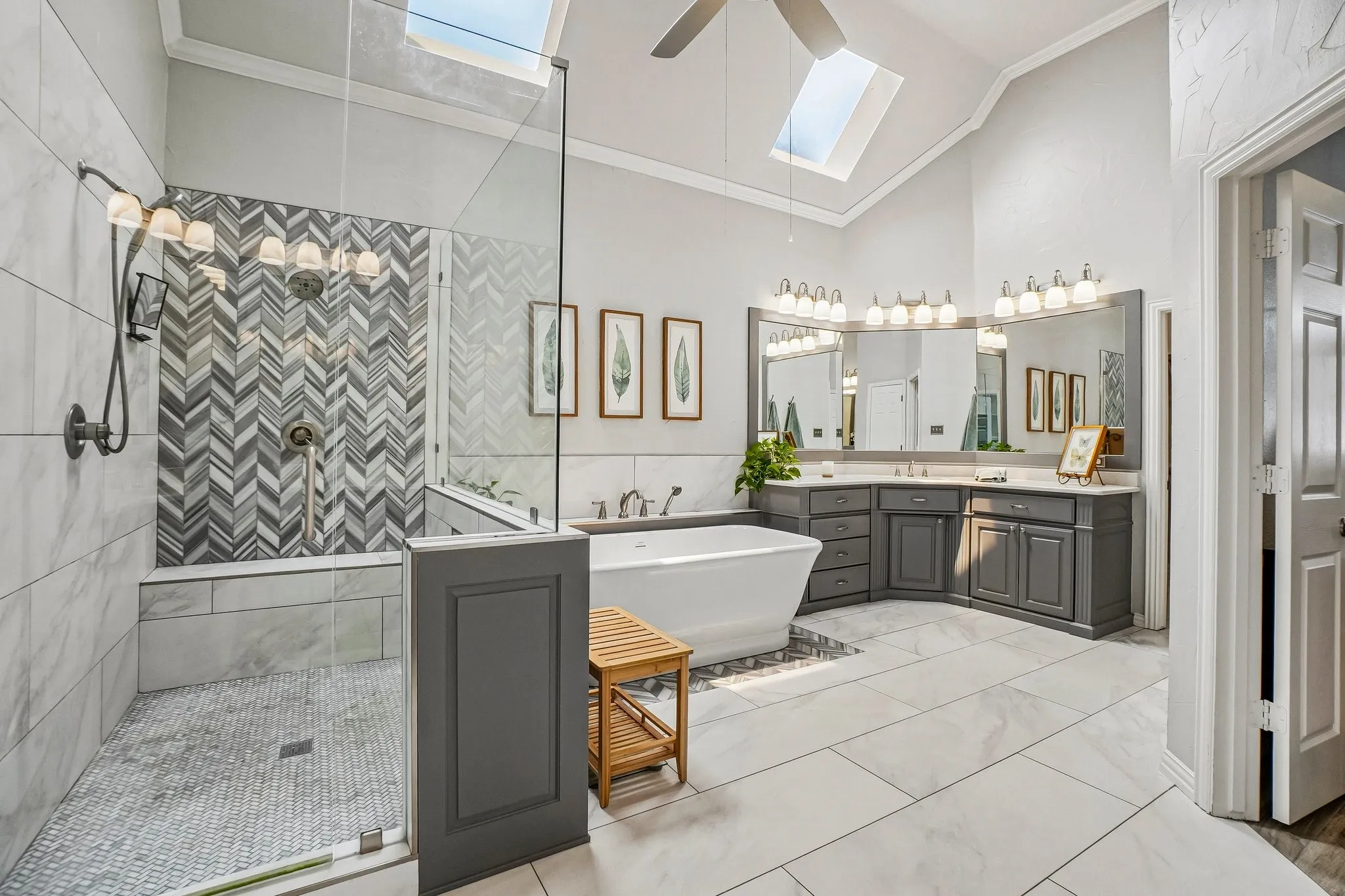 Vaulted ceiling bathroom featuring dual skylights, a glass-enclosed shower with chevron-patterned tile, a freestanding soaking tub, and a dual vanity with grey cabinetry and undermount sinks