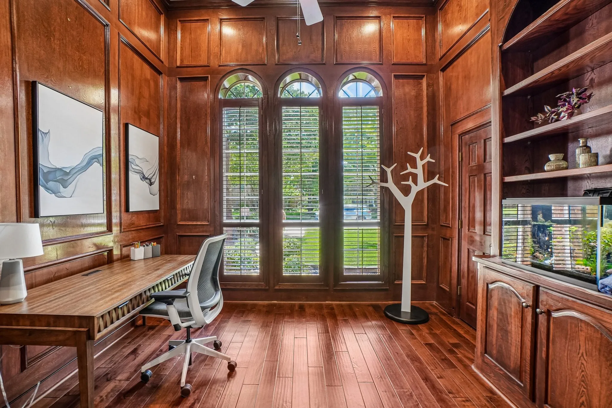 Wood-paneled room featuring three arched windows with plantation shutters, extensive built-in shelving, wood-finish flooring, and a storage closet