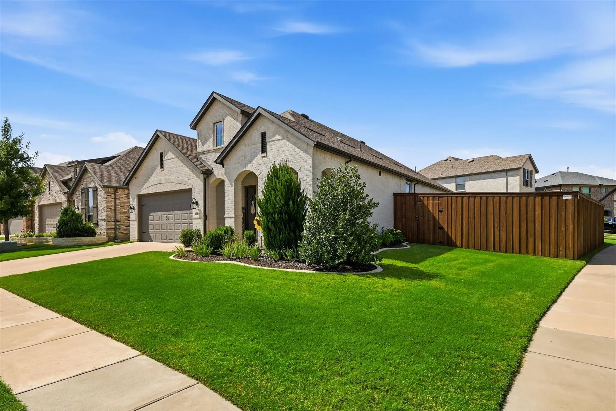 Contemporary stucco and brick exterior featuring a two-car garage, arched entry, mature landscaping, and a manicured lawn