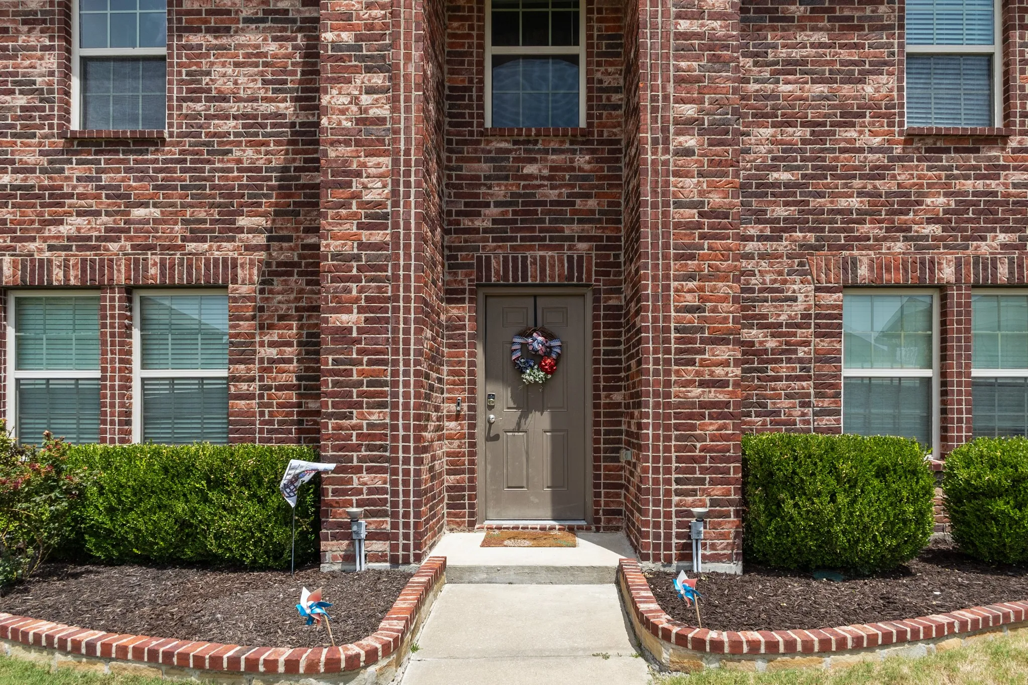 Brick facade home featuring a prominent entryway, a concrete pathway, landscaped beds with brick edging, and multiple windows with blinds