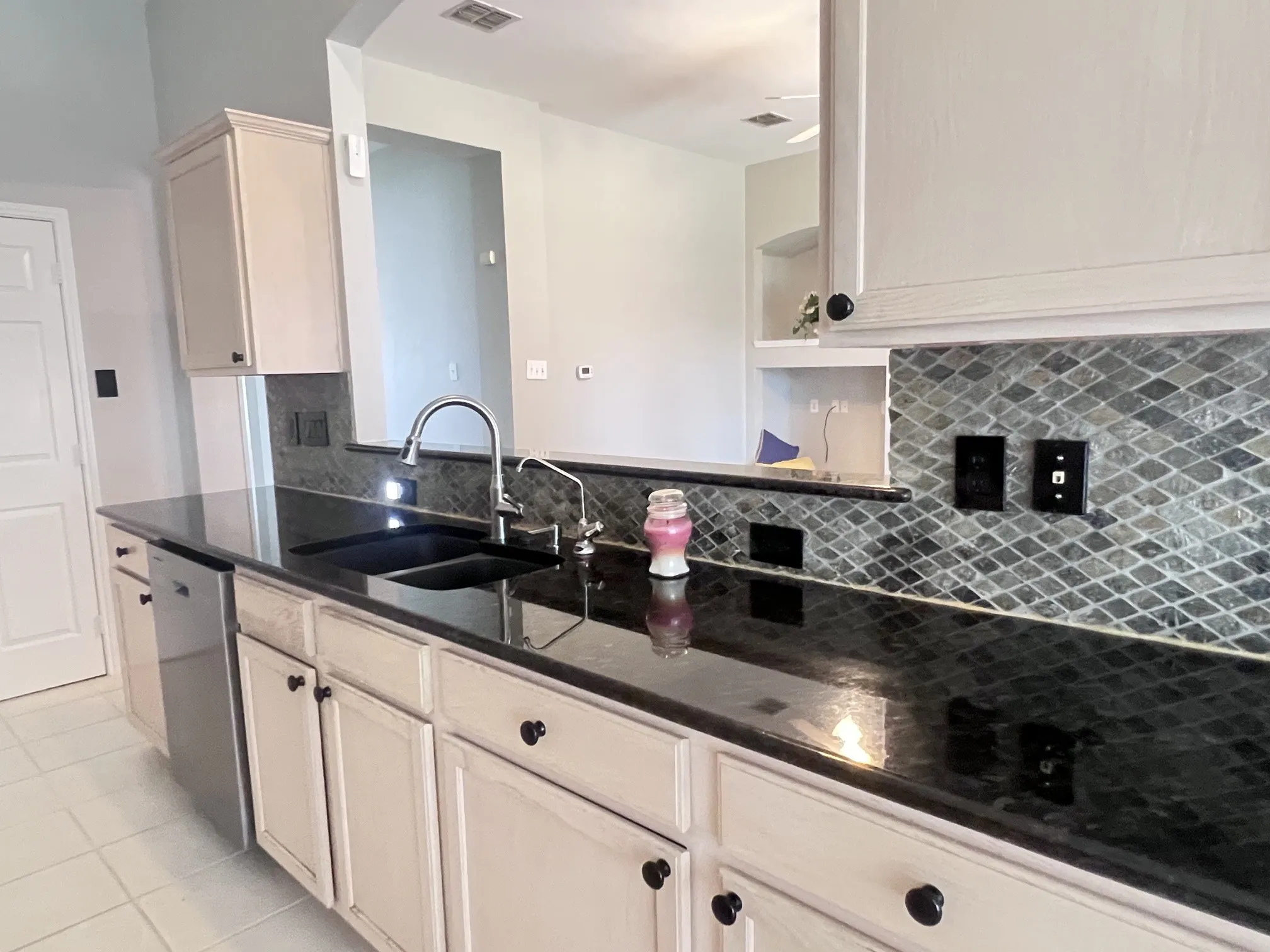 Kitchen featuring a dark stone countertop, light-colored cabinetry with dark hardware, a stainless steel dishwasher, and a tile backsplash with a diamond pattern