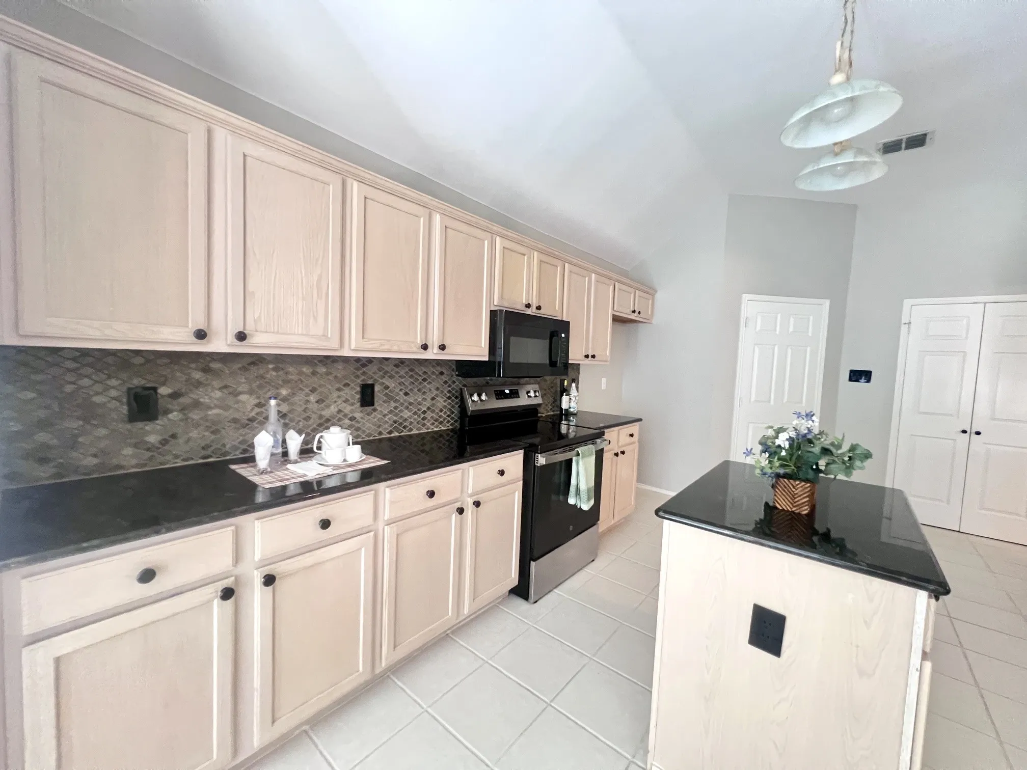Spacious kitchen featuring light wood-finish cabinetry with dark hardware, dark countertops, a patterned tile backsplash, stainless steel appliances, and a central island with a dark countertop