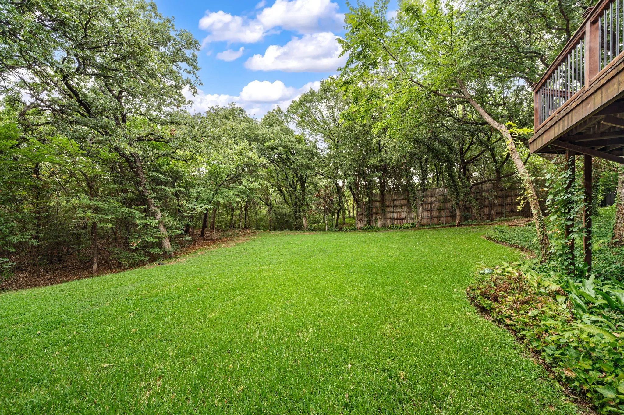Spacious green lawn surrounded by mature trees, featuring a wooden deck with railing and natural landscaping