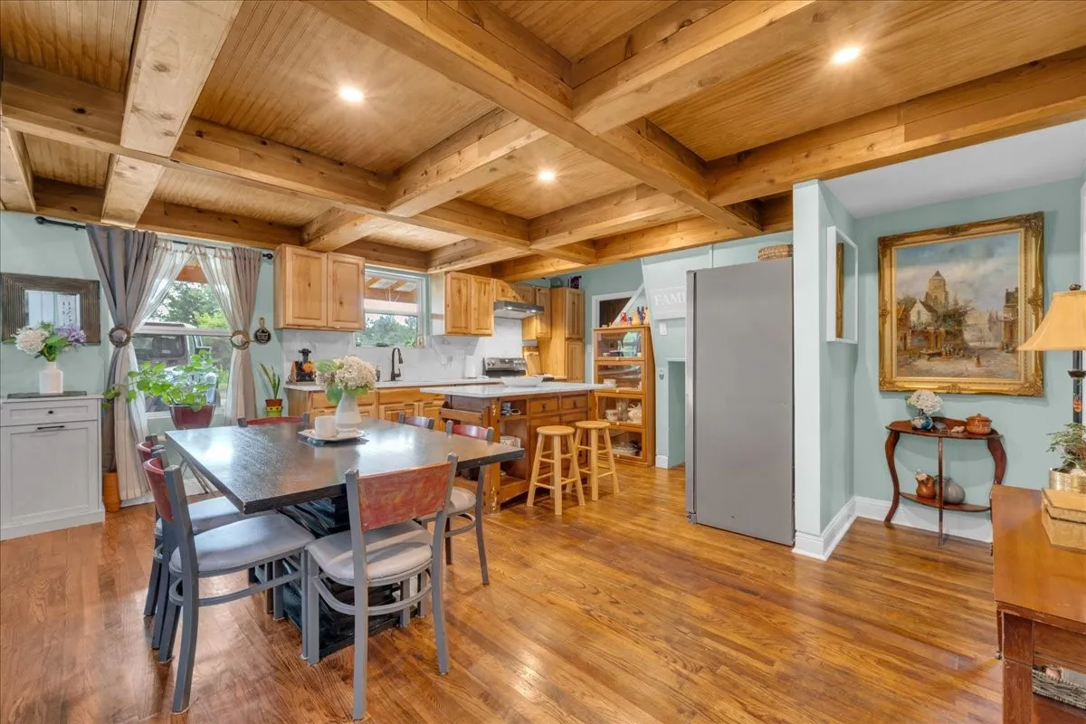 Open-concept kitchen and dining area featuring wood-finish flooring, exposed wood beam ceiling, light wood cabinetry, and a central island with countertop seating