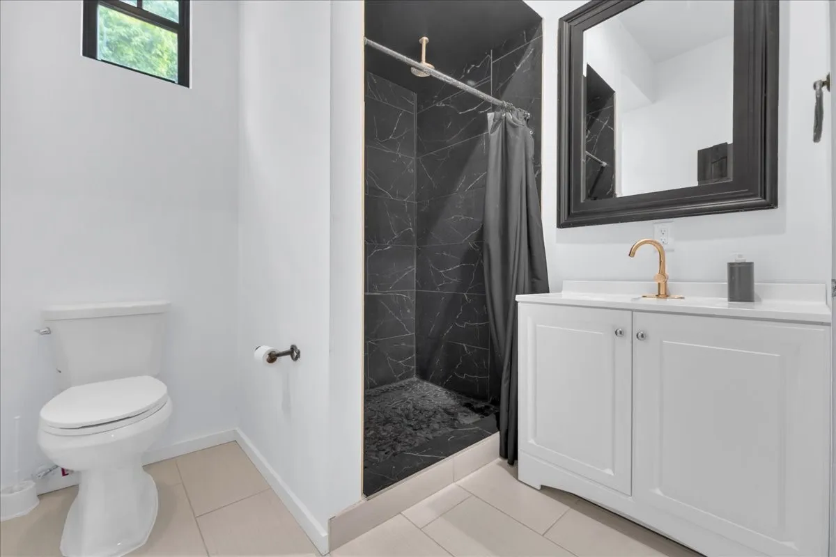 Full bathroom featuring a walk-in shower with black-veined tile, a white vanity with panel doors and gold-finish faucet, and a black framed mirror
