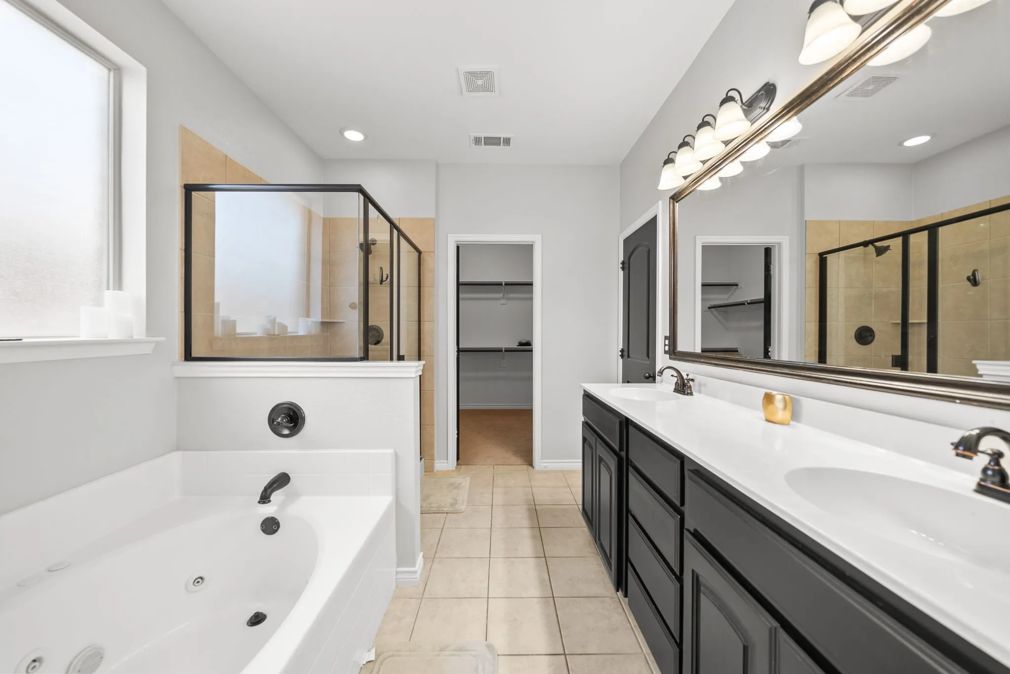 Contemporary bathroom featuring a white soaking tub with dark fixtures, a separate glass-enclosed shower, a double vanity with dark cabinetry and white countertops, and neutral tile flooring