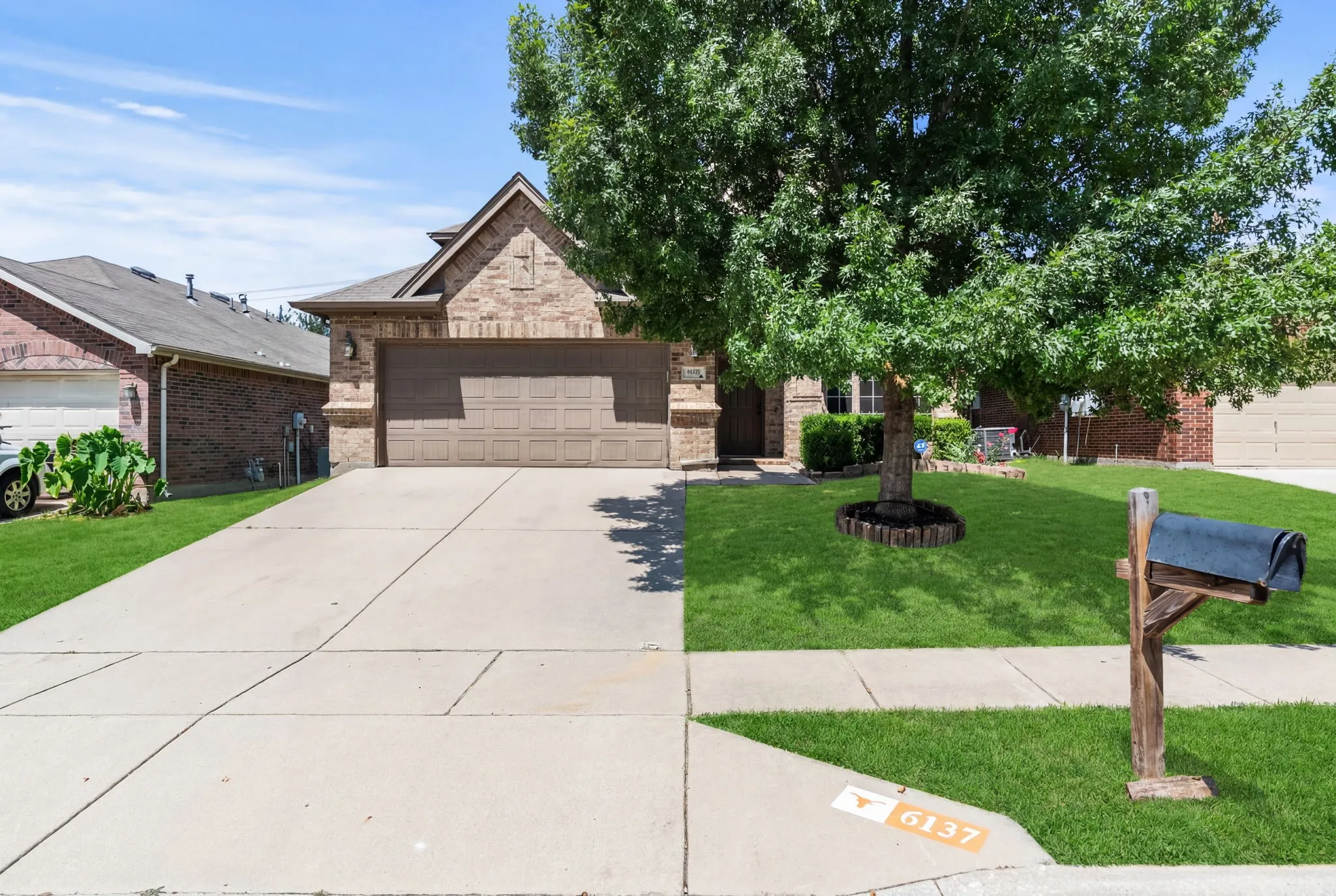 Brick facade residence featuring an attached two-car garage, mature front yard tree, and concrete driveway with sidewalk access