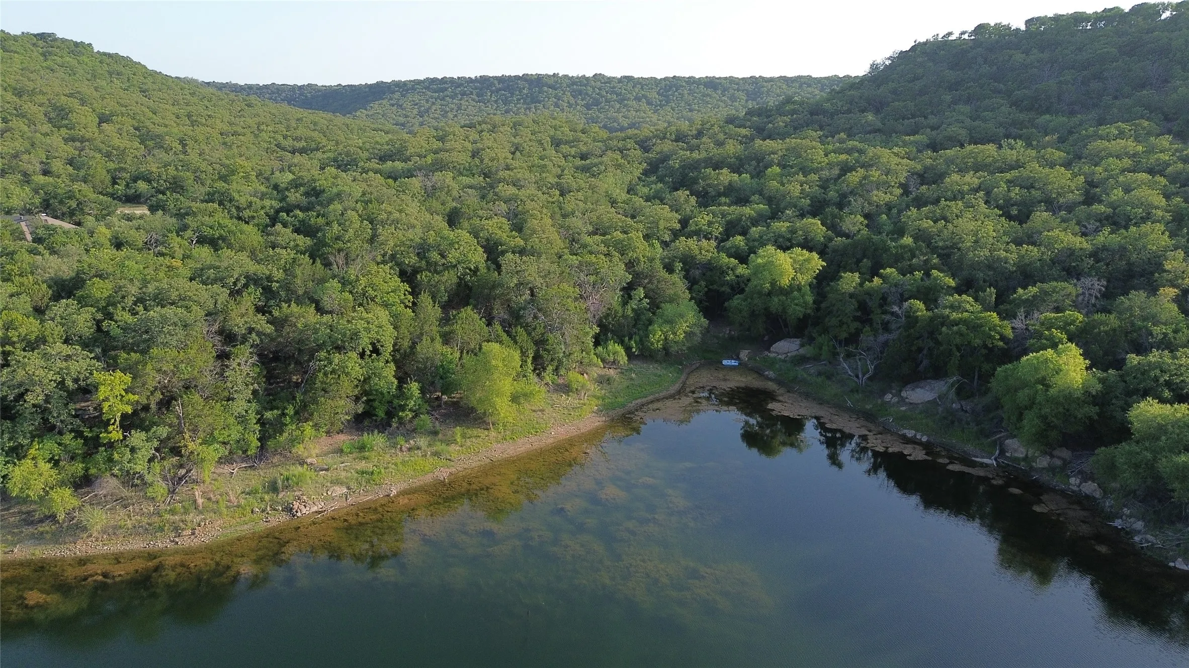 Expansive waterfront property with a verdant tree-lined shoreline, featuring a calm body of water and surrounding wooded hills