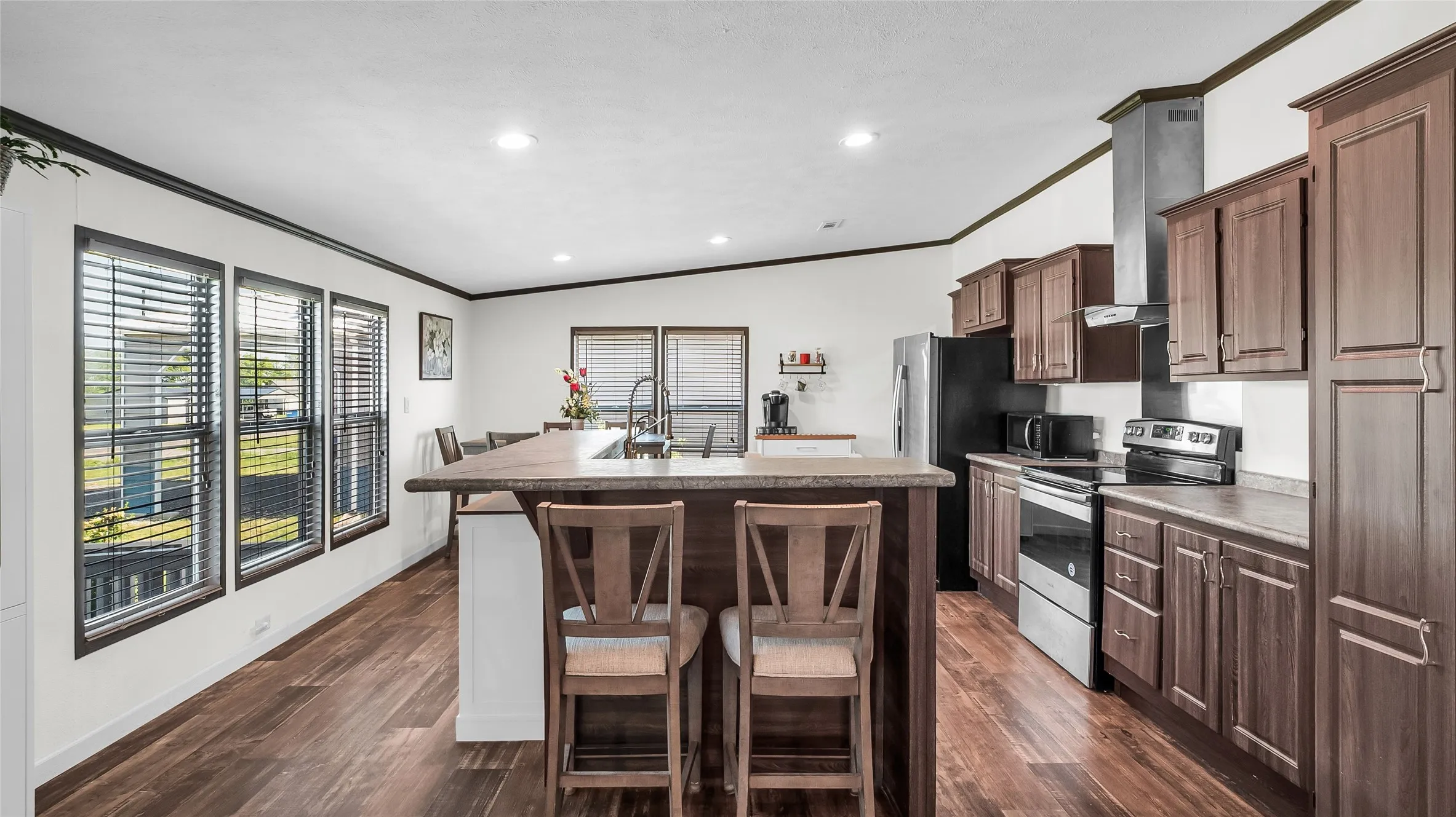 Open-concept kitchen with dark wood cabinetry, a central island with bar seating, and stainless-steel appliances, complemented by recessed lighting and multiple windows with blinds
