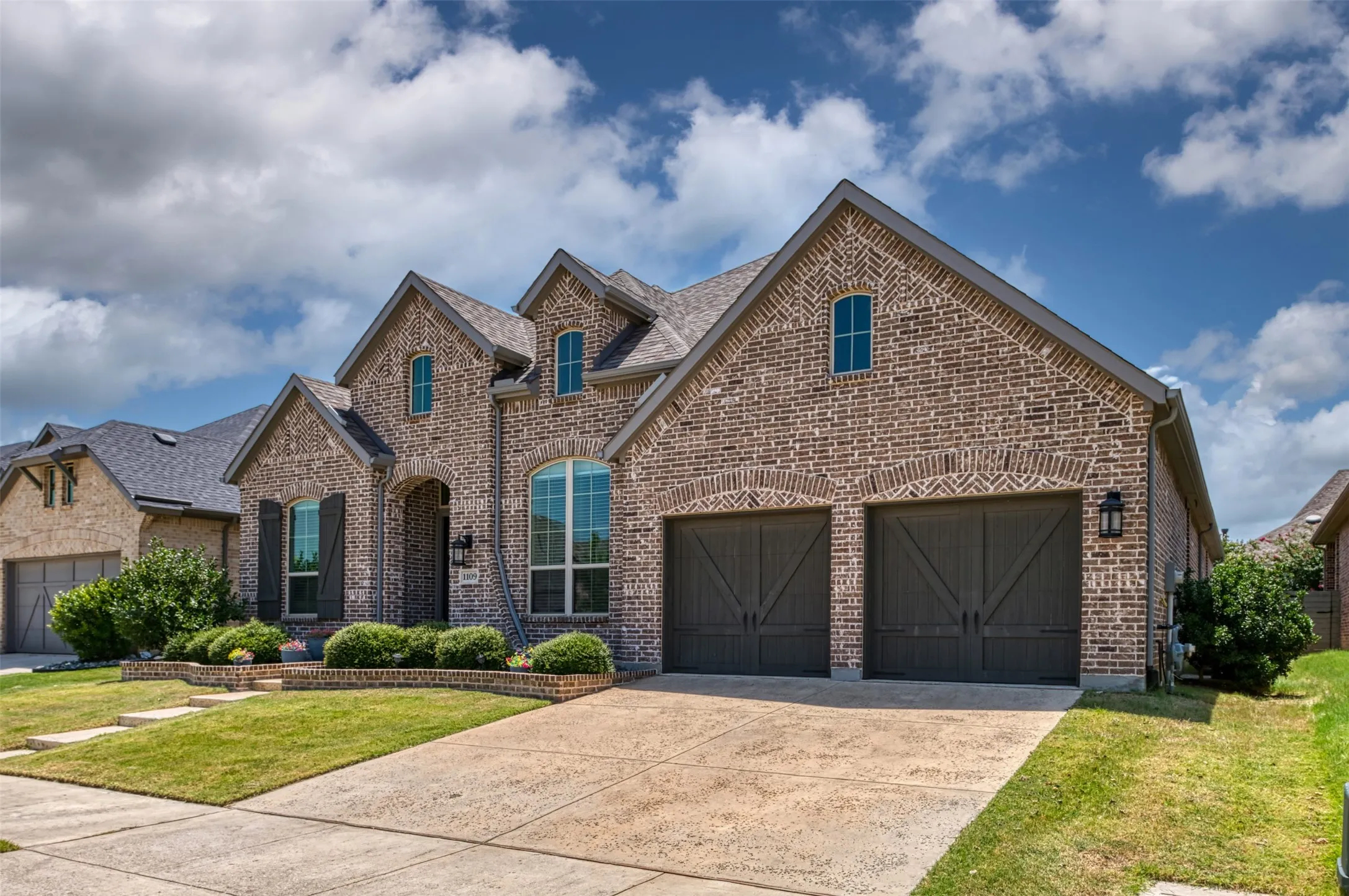Brick exterior featuring a two-car garage with carriage-style doors, arched window accents, gabled rooflines, and a concrete driveway