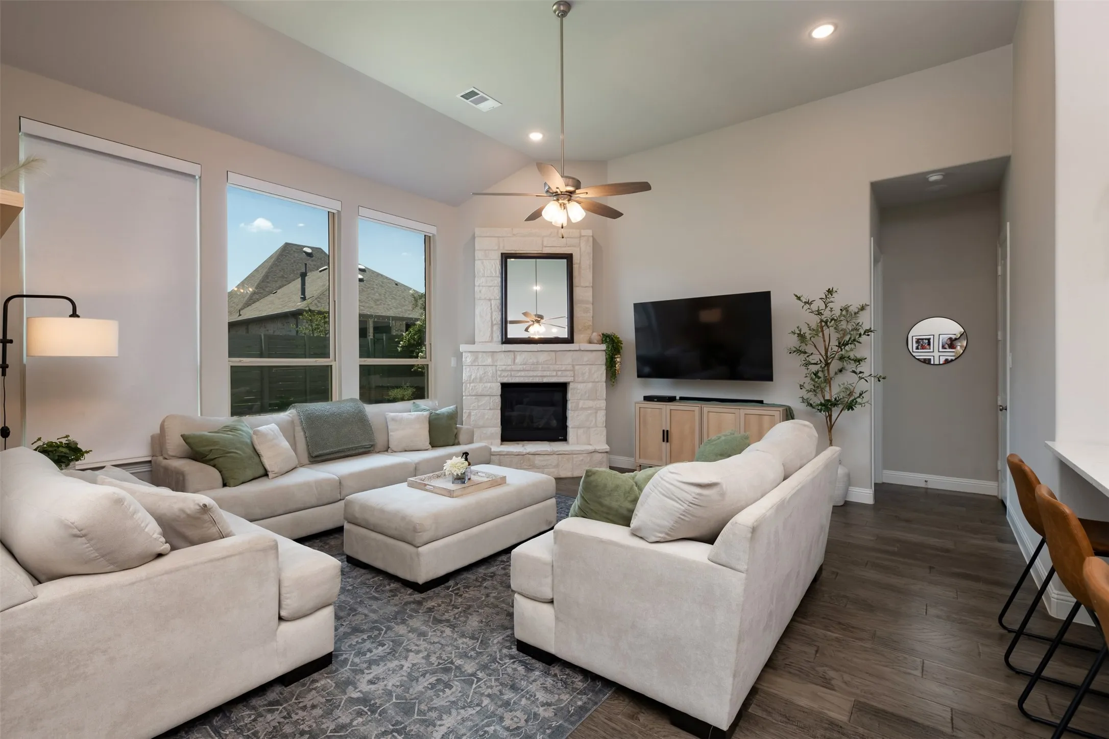 Spacious living area featuring high vaulted ceilings, large windows with roller shades, a stone-clad corner fireplace, and wood-finish flooring