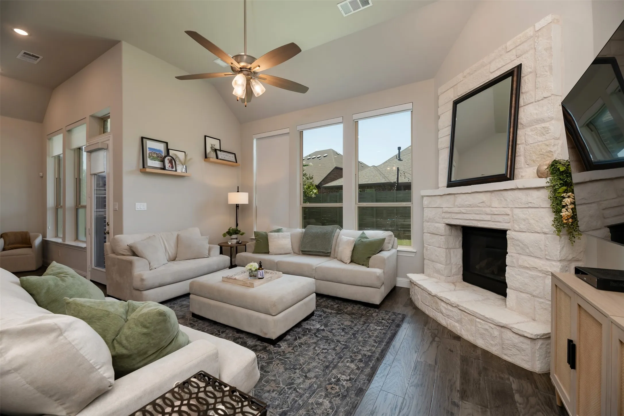 Spacious living area featuring a stone-clad fireplace with hearth, vaulted ceilings, a ceiling fan with integrated lighting, and wood-finish flooring