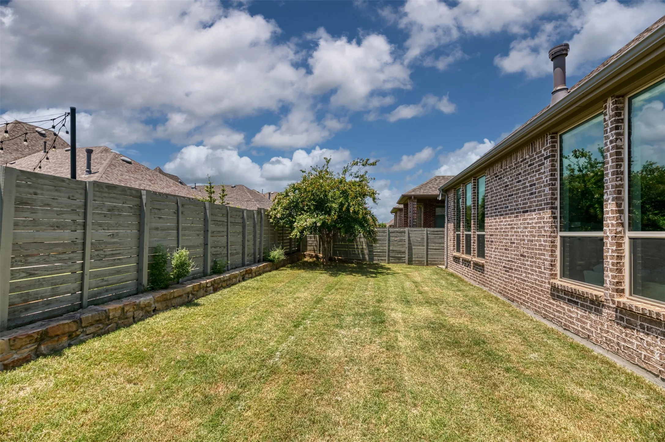 Expansive grassy yard featuring a brick-clad exterior with multiple windows, a horizontal slat fence, a stone retaining wall, and mature landscaping