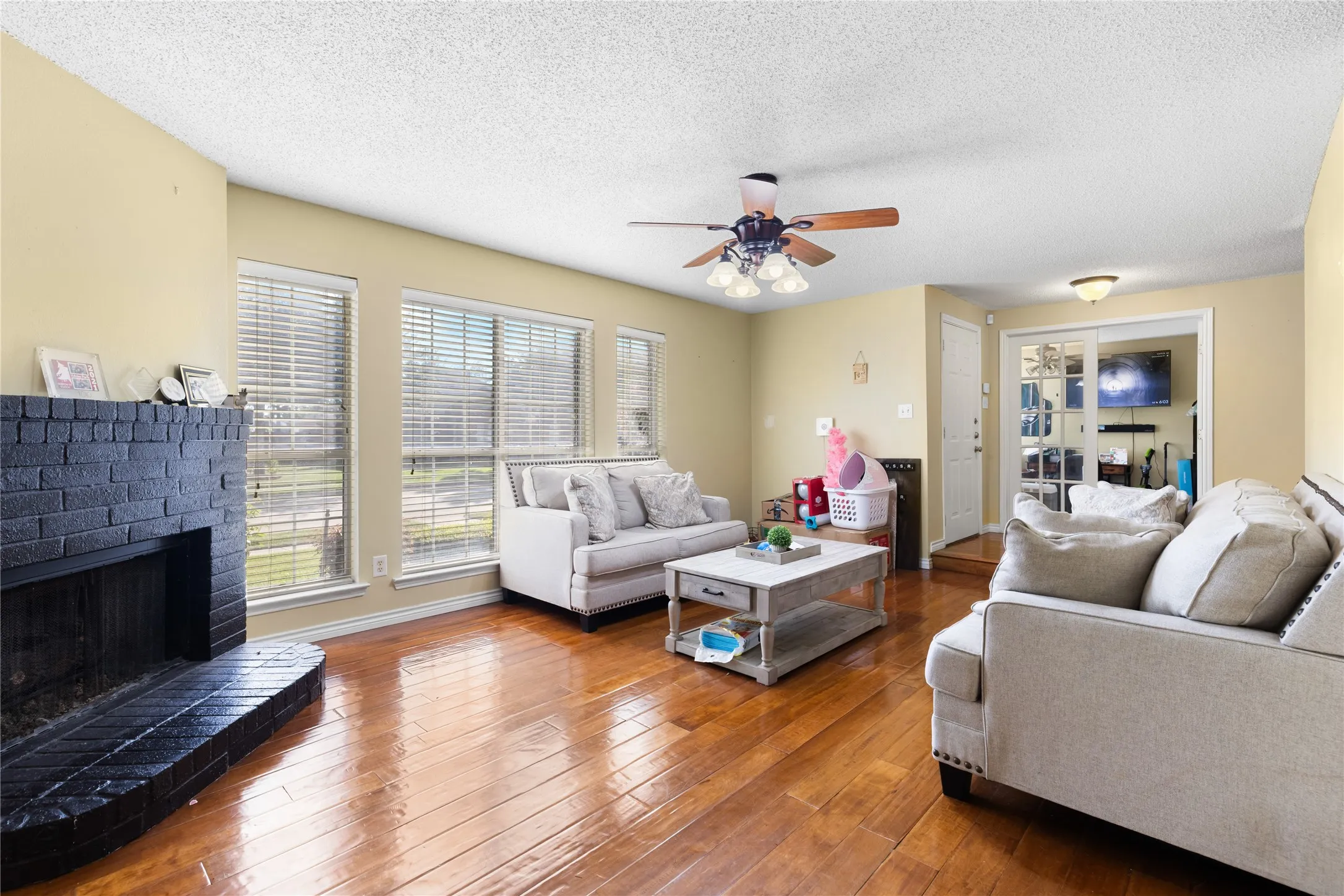 Living area featuring wood-finish flooring, a painted brick fireplace, multiple windows with blinds, a ceiling fan with light fixture, and a doorway with glass-paned French doors