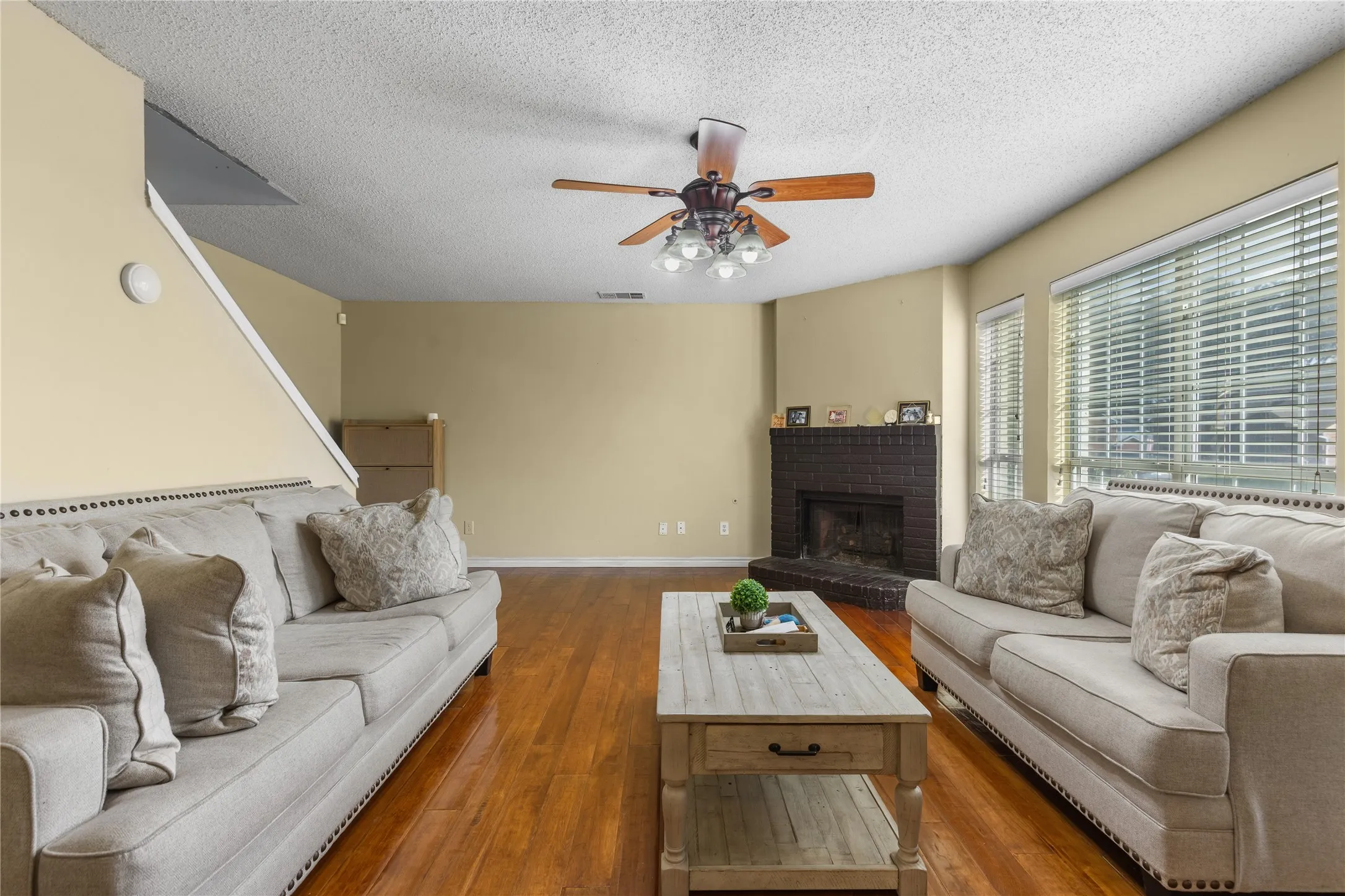 Living space featuring wood-finish flooring, a brick fireplace, a ceiling fan with light kit, and multiple large windows with blinds