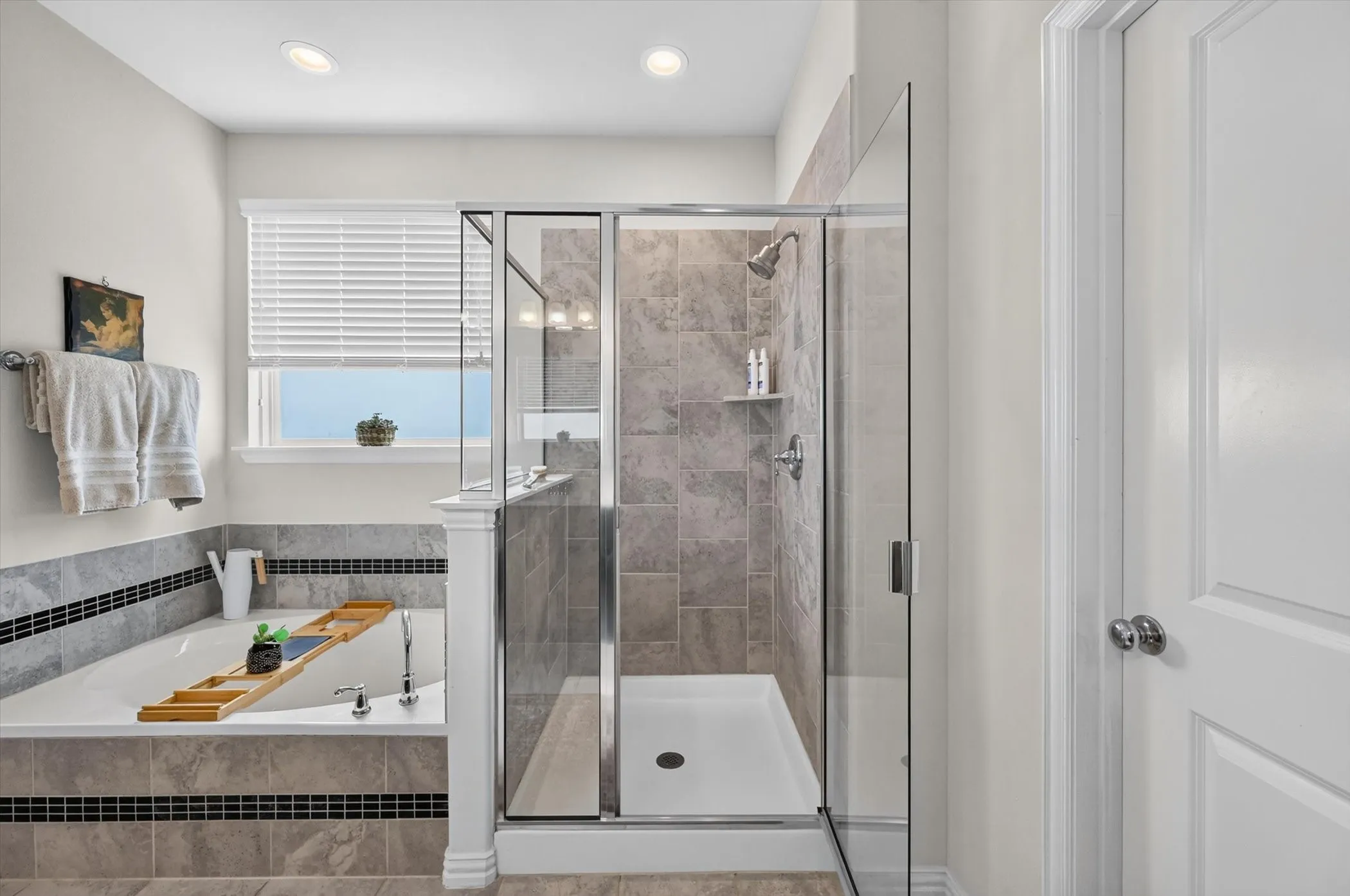 Spacious bathroom featuring a built-in soaking tub with surrounding stone-finish tile, a separate glass-enclosed shower with tiled walls and an integrated shelf, and recessed ceiling lighting
