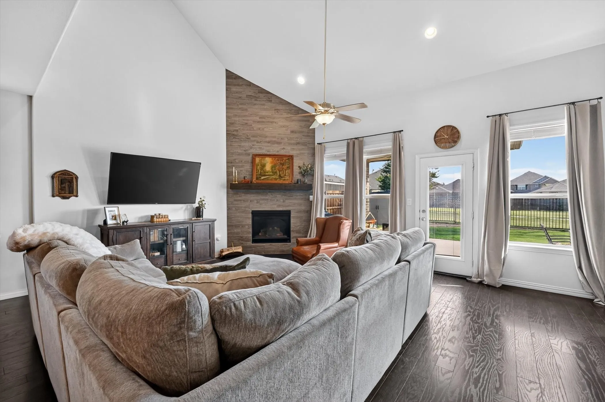 Living area featuring a vaulted ceiling, dark wood-finish flooring, a fireplace with a stone-look surround and wood mantel, and multiple windows with backyard views