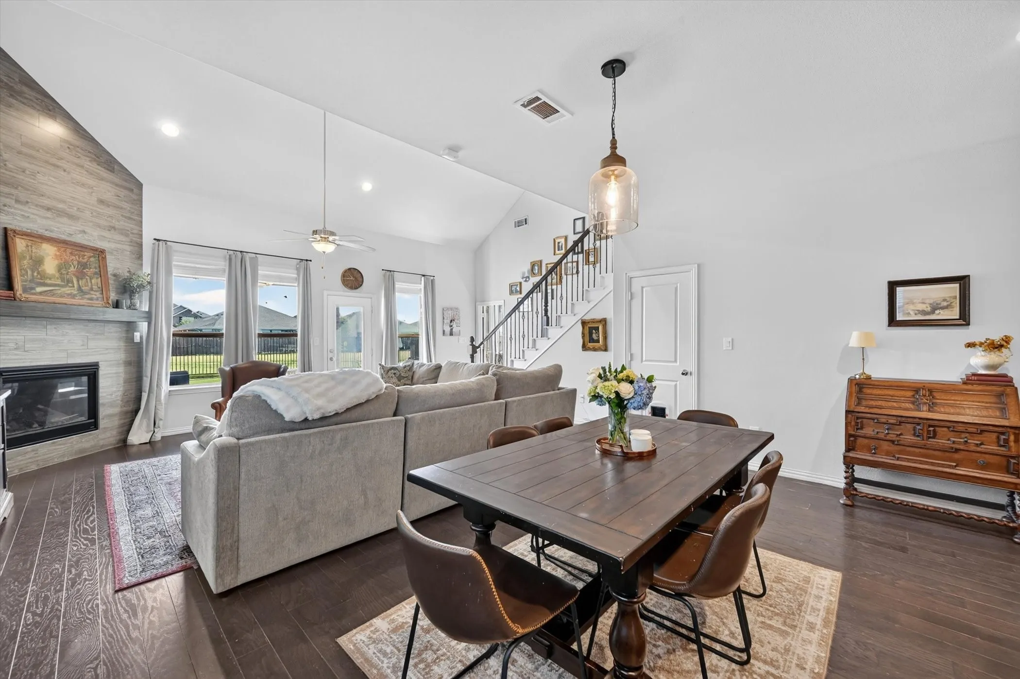 Spacious open concept living area featuring a vaulted ceiling with recessed lighting and a ceiling fan, wood-finish flooring, a contemporary fireplace with a stone surround and wood-paneled accent wall, and a dining area with a distinctive pendant light fixture