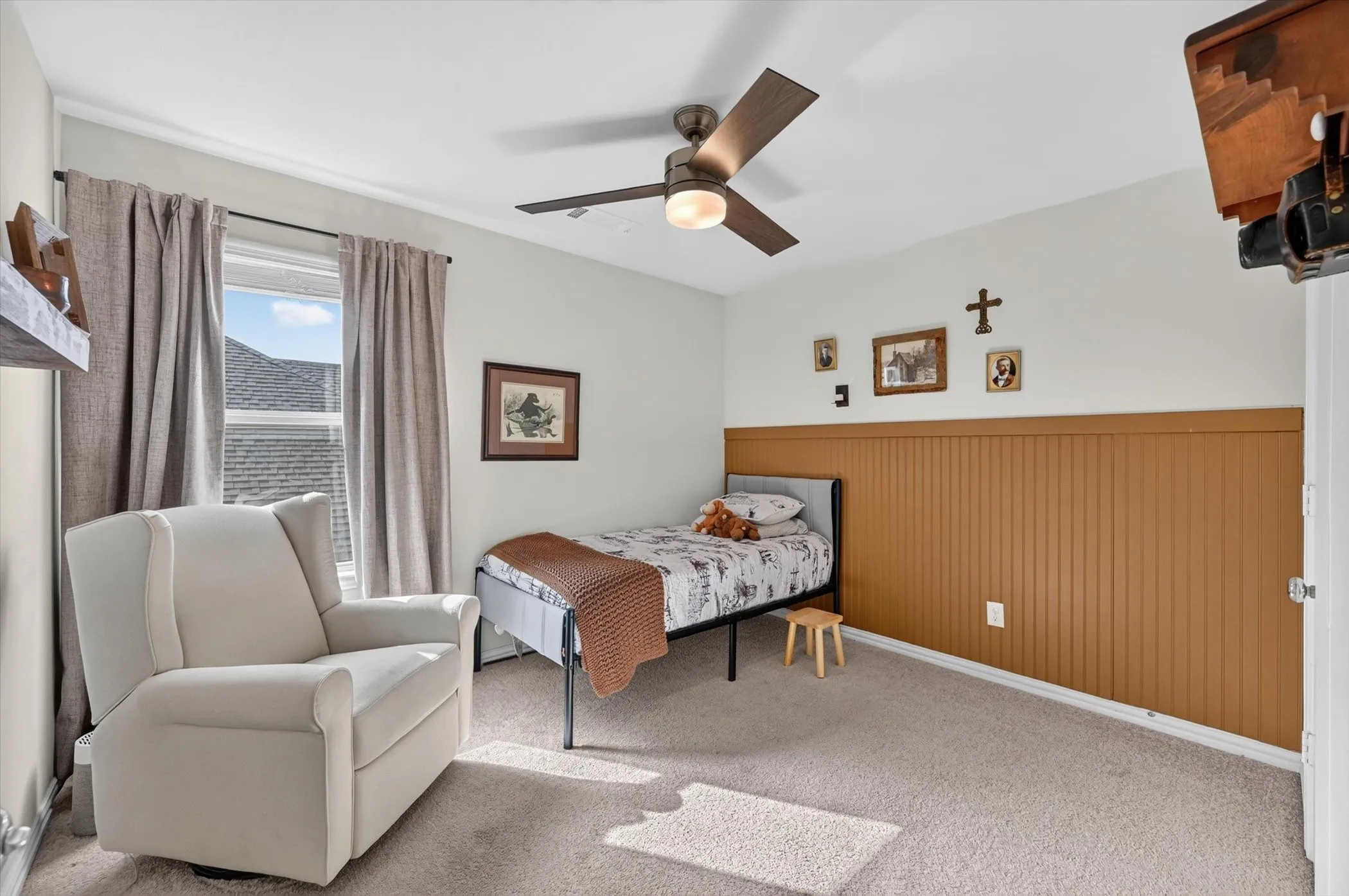 Bedroom featuring light neutral wall paint, wood-finish ceiling fan with integrated lighting, window with drapes, and a wainscoting accent wall