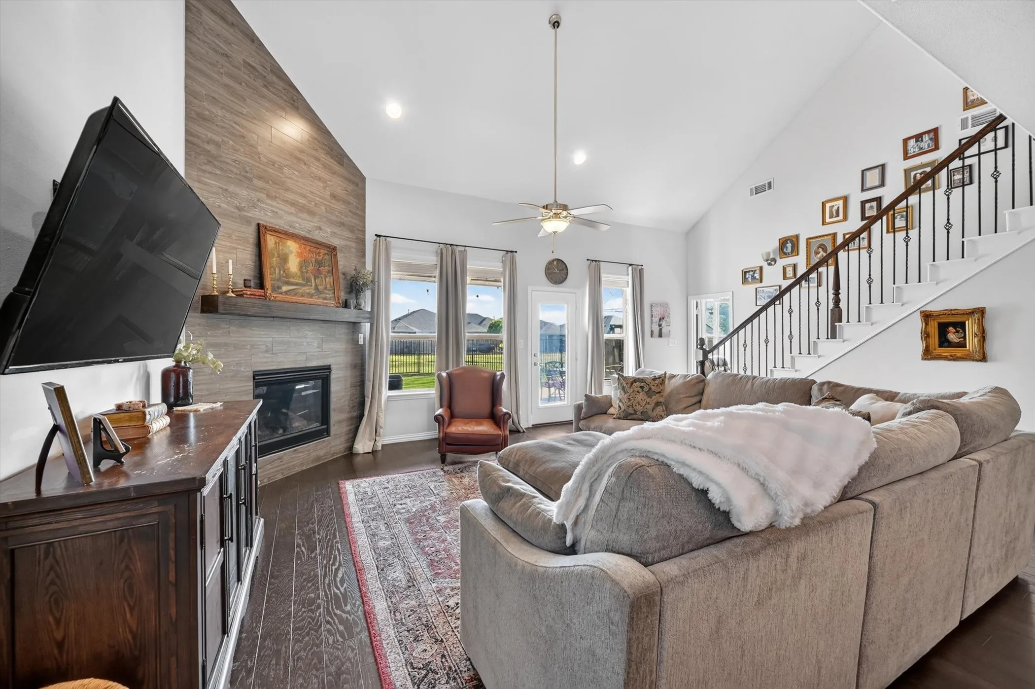Living area with vaulted ceiling, wood-finish flooring, a contemporary fireplace with a timber mantel, a multi-panel glass door to the exterior, and an open staircase with wrought iron balusters