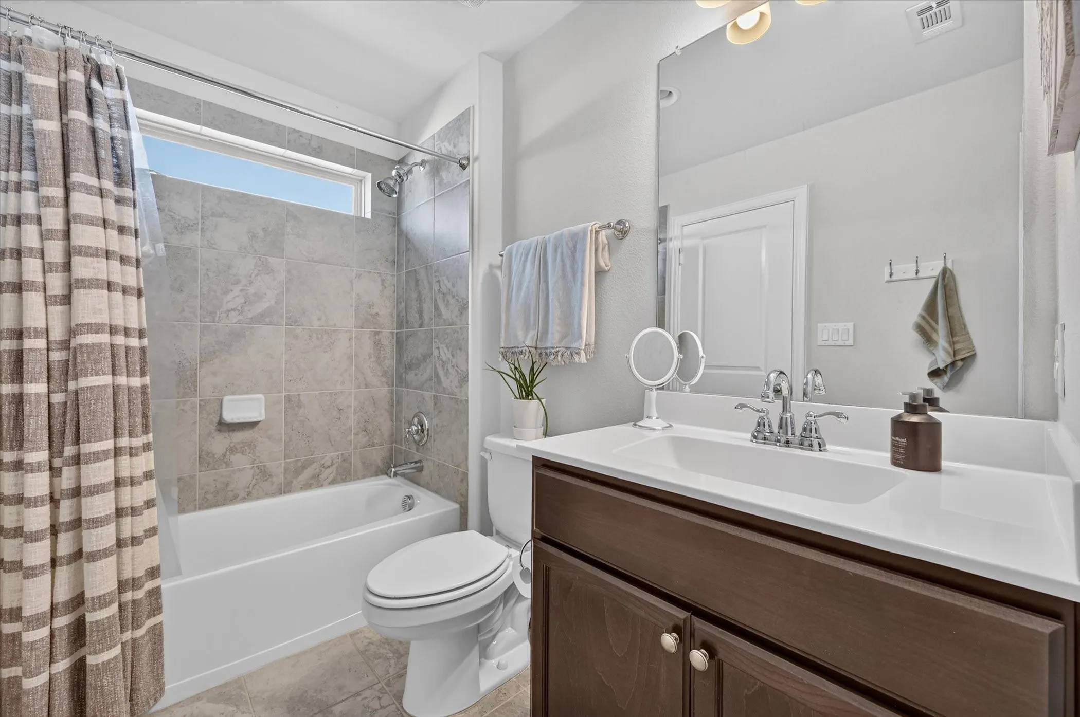 Bathroom featuring a tiled tub-shower combination with a horizontal window, a white toilet, a dark wood-finish vanity with a white countertop and integrated sink, and a large wall-mounted mirror